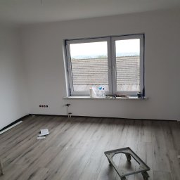 Gdzbouw - Pokój po remoncie z panelami podłogowymi i świeżo pomalowanymi ścianami na biało. Widoczne narzędzia i drabina. Duże okno z widokiem na dachy.
