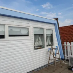 Biała elewacja domu z oknami i niebieskimi elementami wykończeniowymi, drabina malarska oparta o ścianę, widok na błękitne niebo i czerwony dach sąsiedniego budynku.