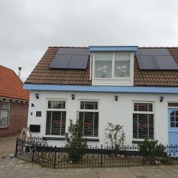 Gdzbouw - Biały dom z niebieskimi akcentami i panelami słonecznymi na brązowym dachu, widziany z poziomu ulicy. W tle widoczny sąsiedni dom z czerwonej cegły.