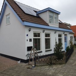 Gdzbouw - Biały dom z niebieskimi akcentami po renowacji elewacji w Brzegu. Widoczne panele słoneczne na dachu, drabina i akcesoria malarskie przy ścianie.