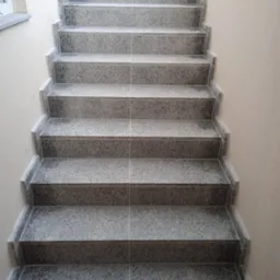 Wykończone schody zewnętrzne z szarego granitu, widok z dołu. Estetyczne połączenia płytek, jasne ściany, delikatne oświetlenie. Precyzyjne wykonanie.