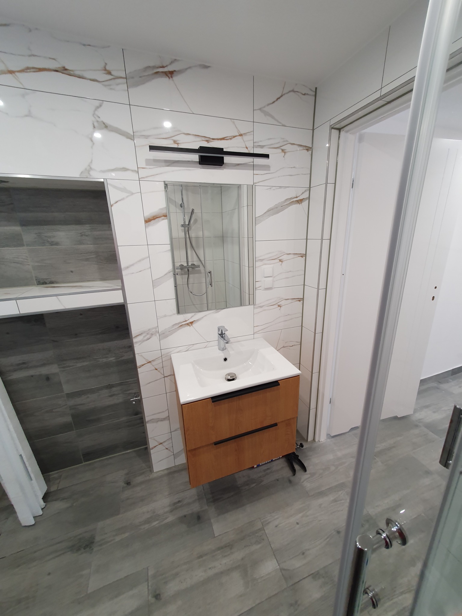 Nowoczesna łazienka po remoncie: umywalka z szafką z drewna, białe płytki imitujące marmur z złotymi żyłami na ścianach, szare płytki podłogowe imitujące drewno, kabina prysznicowa ze szklanymi...