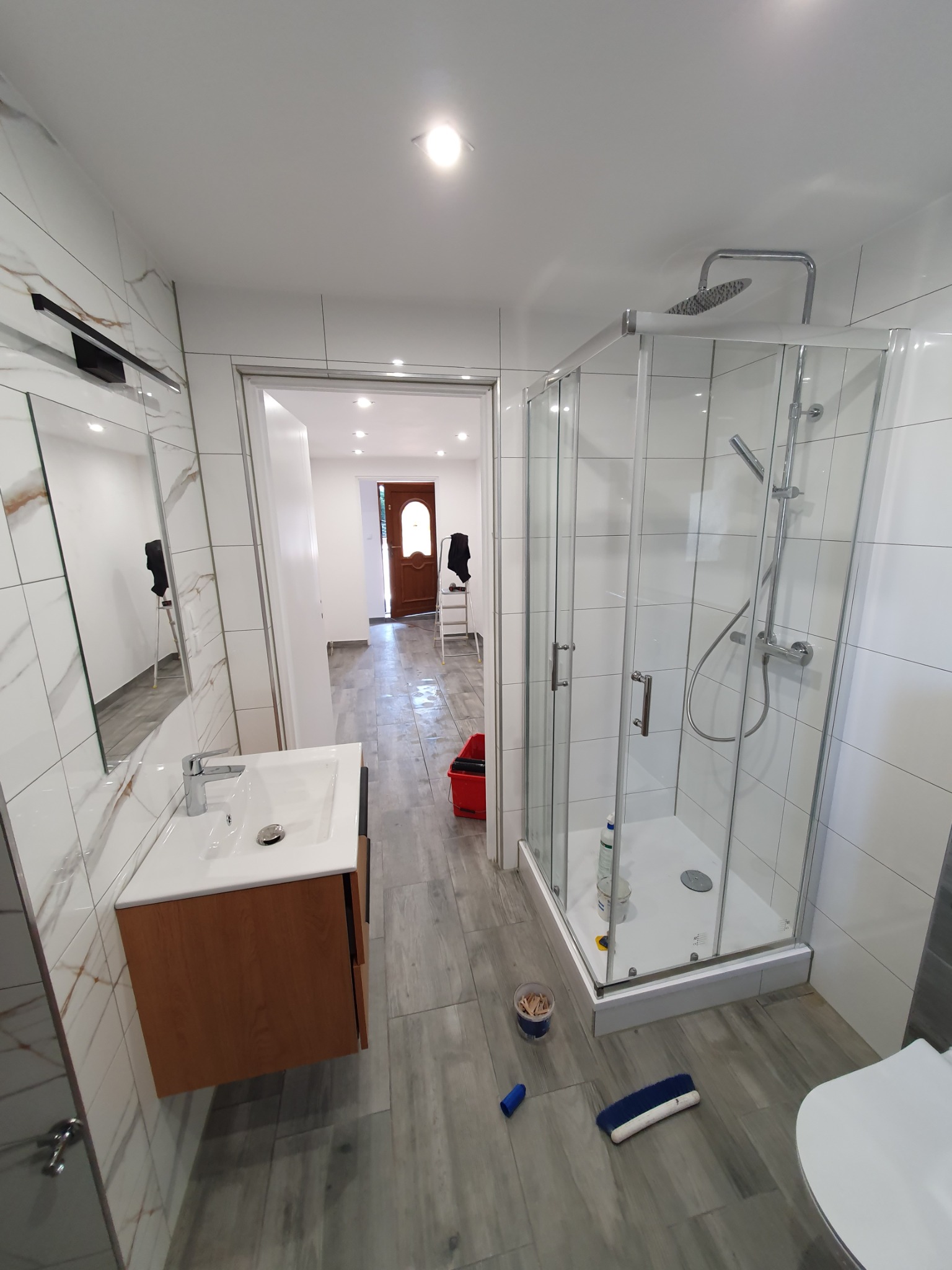 Wykończona łazienka z kabiną prysznicową, umywalką z szafką w kolorze drewna, płytkami imitującymi marmur na ścianach i szarymi płytkami podłogowymi. W tle widoczny otwarty pokój z czerwoną...