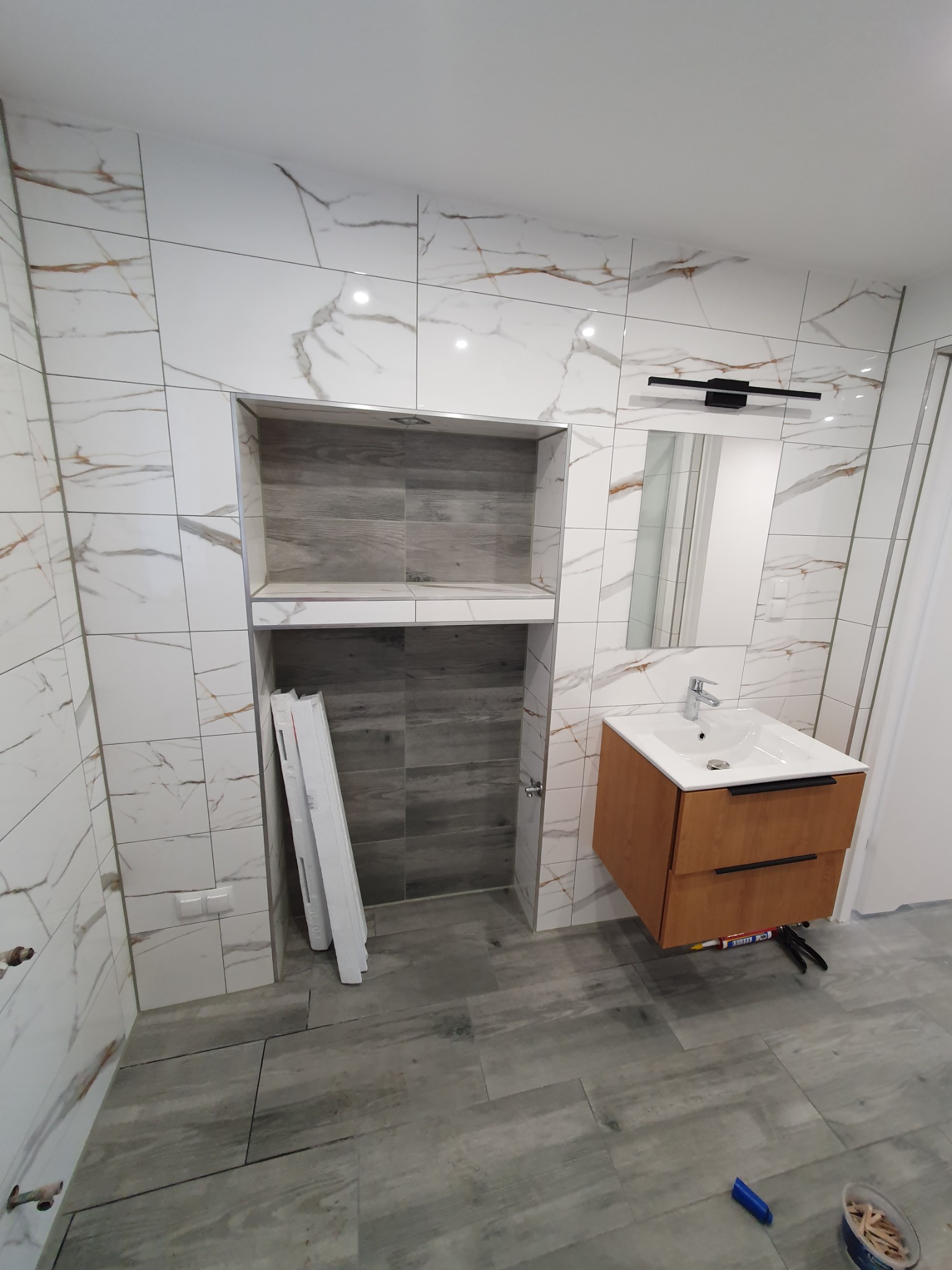 Wykończona łazienka z marmurowymi płytkami ściennymi, drewnianą szafką pod umywalką i wnęką w ścianie wyłożoną szarymi płytkami imitującymi drewno. Na podłodze szare płytki. We wnęce leżą białe...