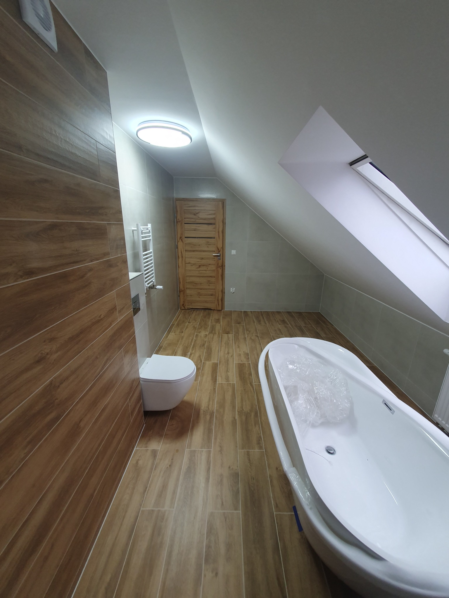 Jasna łazienka na poddaszu z drewnopodobnymi płytkami, białą wanną wolnostojącą i toaletą. Widoczne okno dachowe i oświetlenie sufitowe.