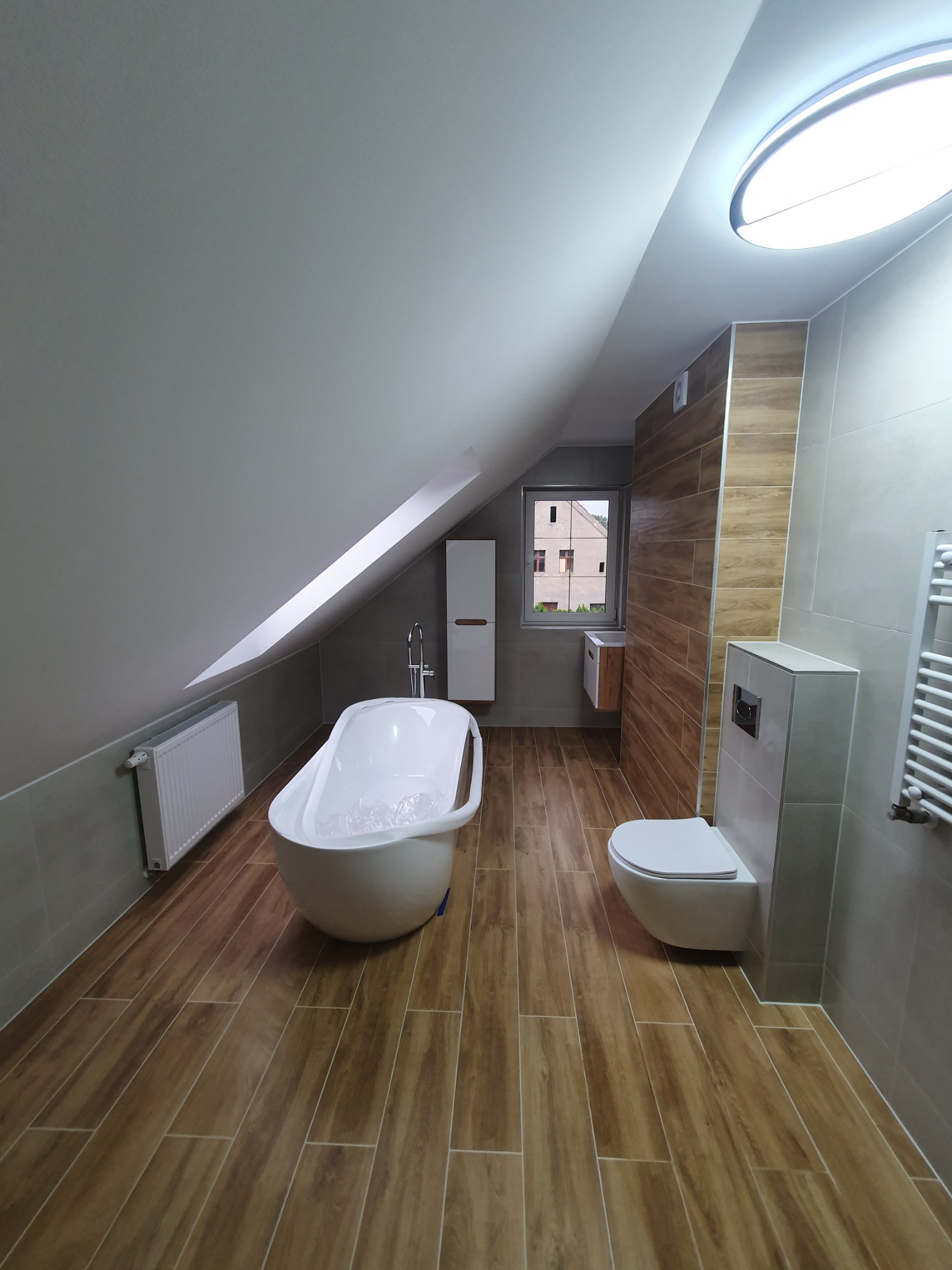Nowoczesna łazienka na poddaszu z wolnostojącą wanną, drewnopodobną podłogą i szarą glazurą. Uwagę zwraca okno z widokiem na budynek.