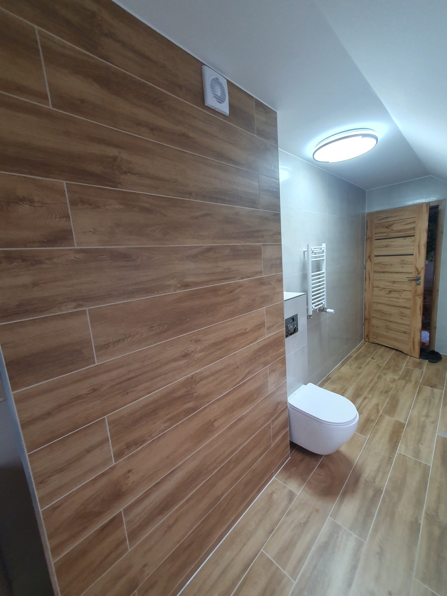 Nowoczesna łazienka po remoncie z drewnopodobnymi płytkami na ścianach i podłodze, wiszącą toaletą i grzejnikiem drabinkowym. Widoczny wentylator.