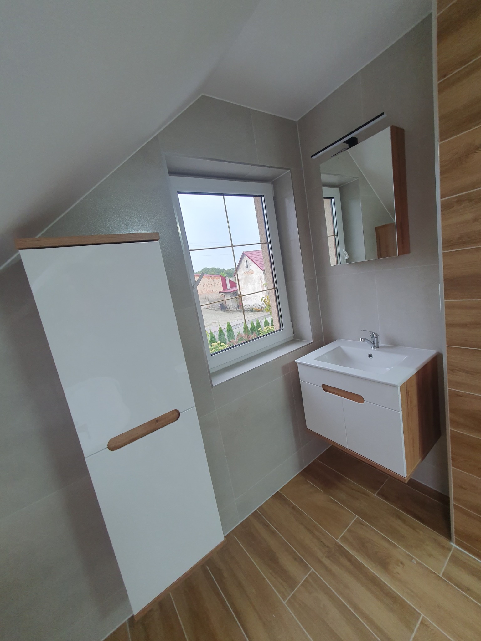 Jasna łazienka na poddaszu z oknem: szare płytki, drewnopodobna podłoga, białe szafki z drewnianymi uchwytami, lustro z oświetleniem. Widok na zielony ogród za oknem.