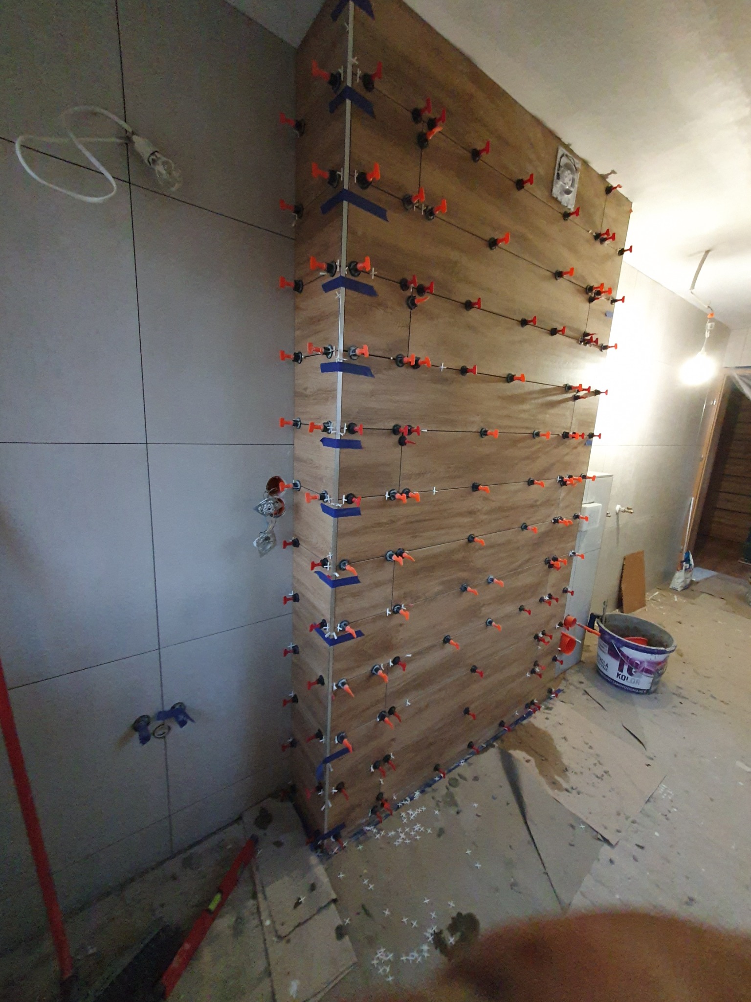 Ściana z płytek imitujących drewno, w trakcie montażu z użyciem systemu poziomowania. Widoczne krzyżyki i kliny. Częściowo ułożone płytki na sąsiedniej ścianie.