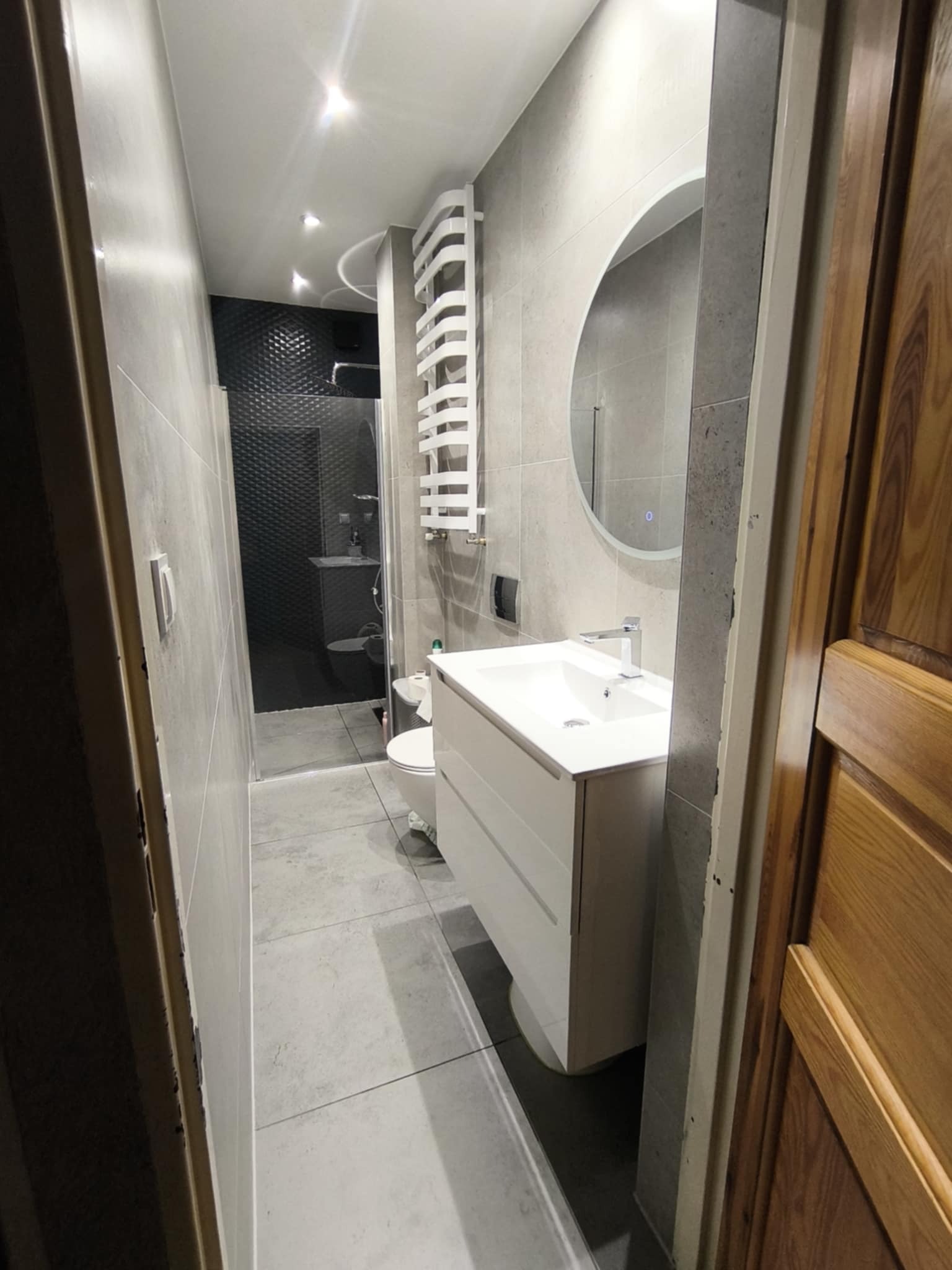 Nowoczesna łazienka z szarą płytką, owalnym lustrem z podświetleniem i białymi meblami. Widoczny prysznic z czarną mozaiką i grzejnik drabinkowy.