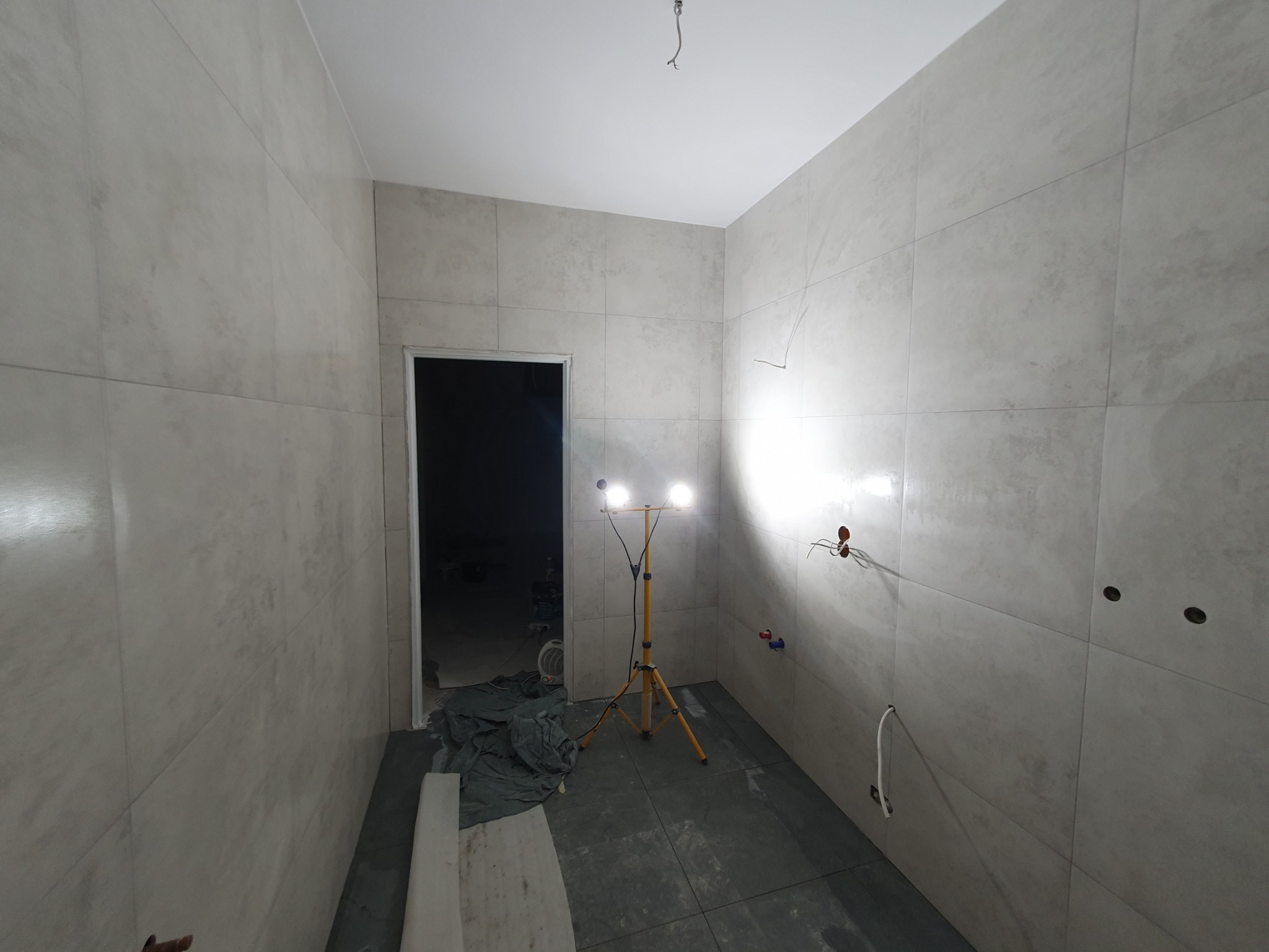 Wykończona łazienka z beżowymi płytkami, widoczne otwory na instalacje, podłoga wyłożona ciemnymi płytkami, oświetlenie budowlane. W tle otwarte drzwi.