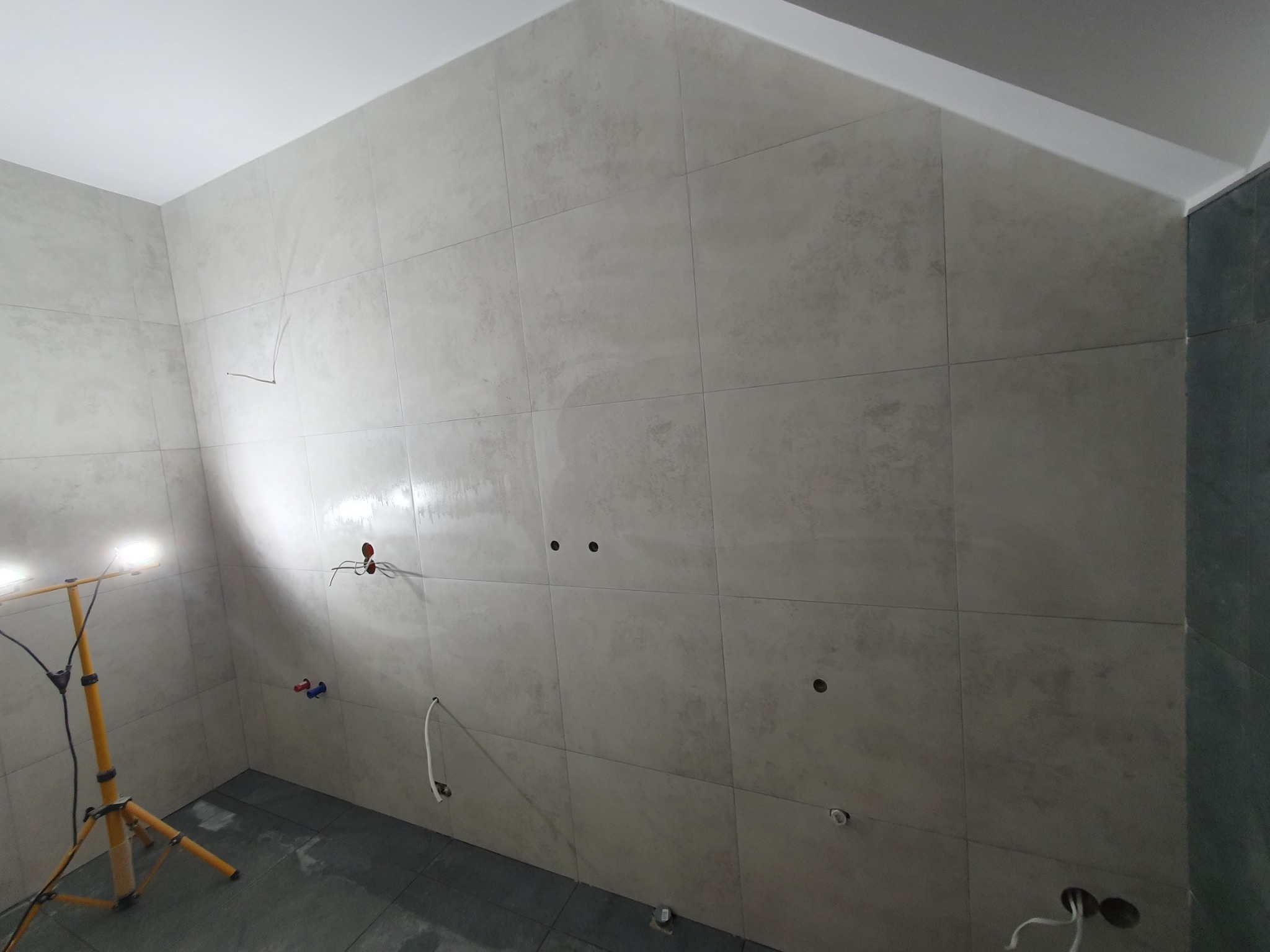 Ściana w łazience po ułożeniu szarych płytek, widoczne otwory na instalacje, oświetlenie budowlane. Minimalistyczny styl, surowe wykończenie.