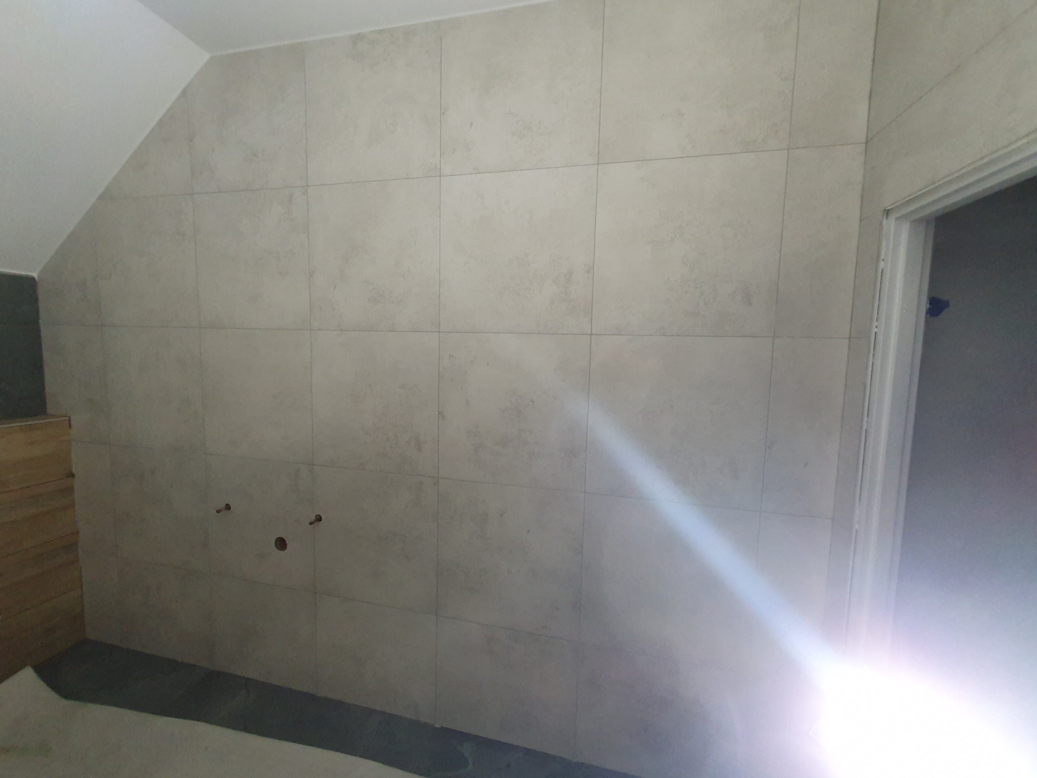 Wykończona ściana w łazience z beżowych, matowych płytek. Widoczne przyłącza wodne i fragment drzwi. Ściana na poddaszu.