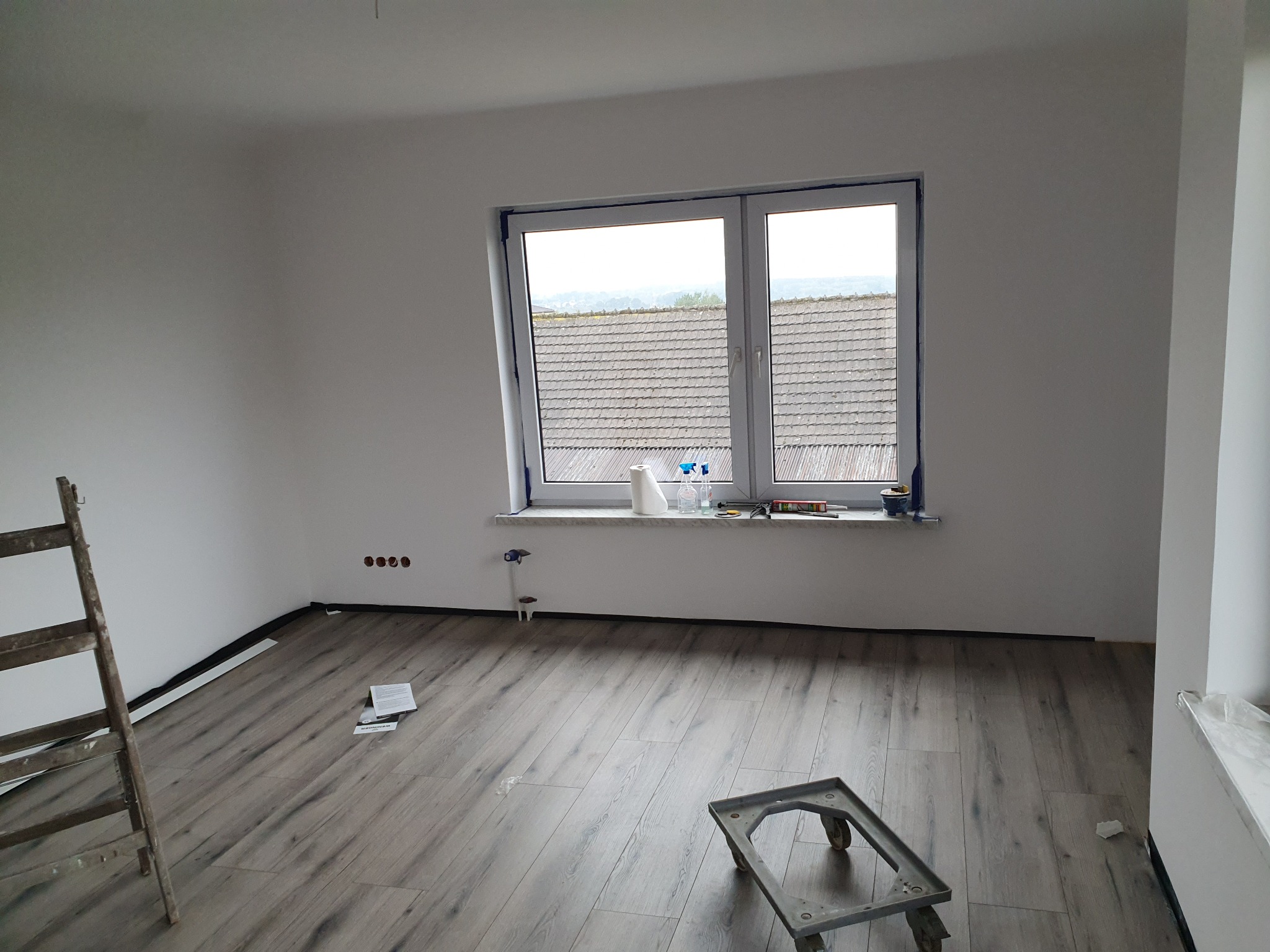 Pokój po remoncie z panelami podłogowymi i świeżo pomalowanymi ścianami na biało. Widoczne narzędzia i drabina. Duże okno z widokiem na dachy.
