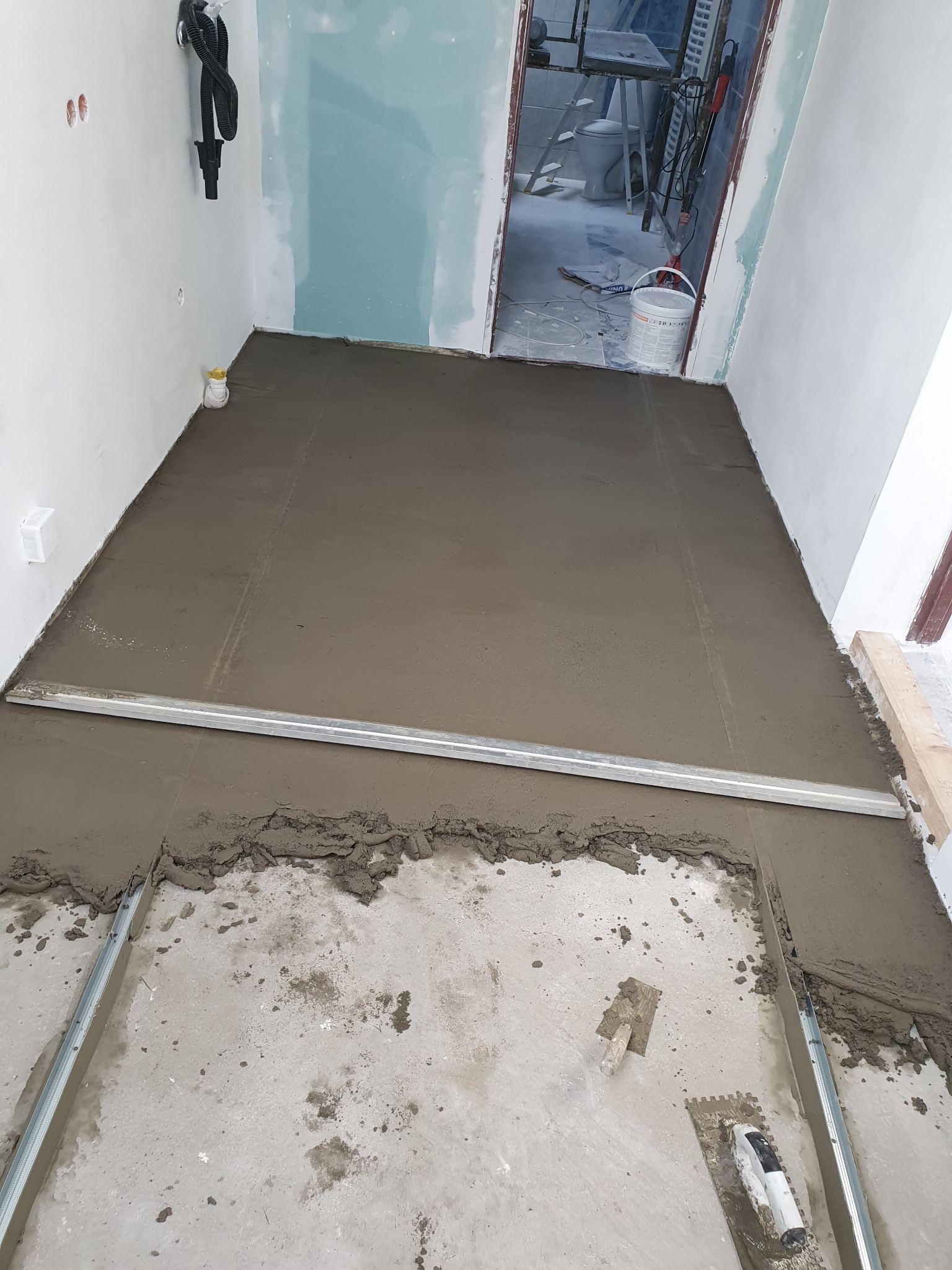 Wyrównywanie podłoża w łazience za pomocą poziomicy i szpachli. Widoczne narzędzia i częściowo ułożona wylewka samopoziomująca. W tle wejście do pomieszczenia.