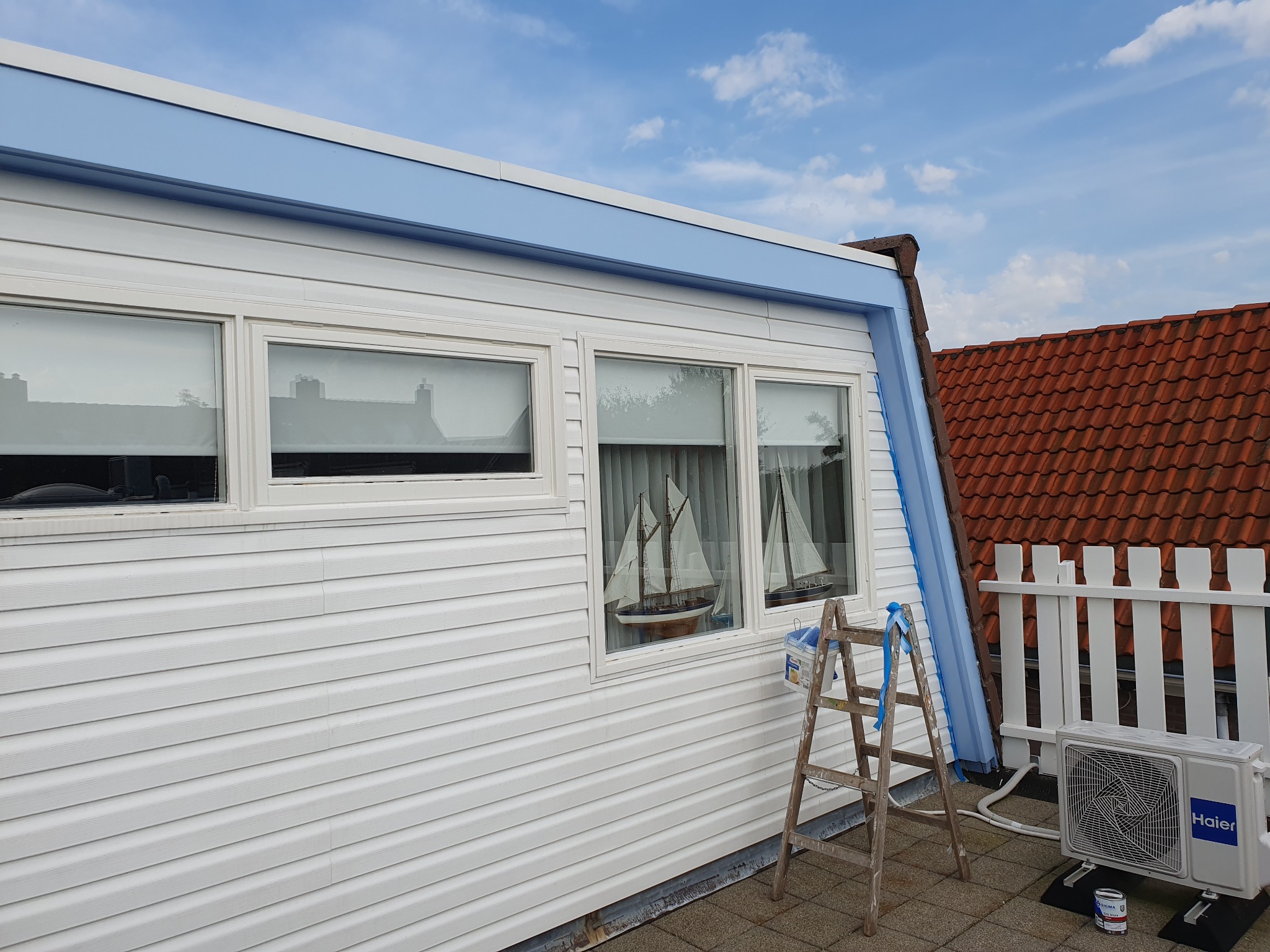 Biała elewacja domu z oknami i niebieskimi elementami wykończeniowymi, drabina malarska oparta o ścianę, widok na błękitne niebo i czerwony dach sąsiedniego budynku.