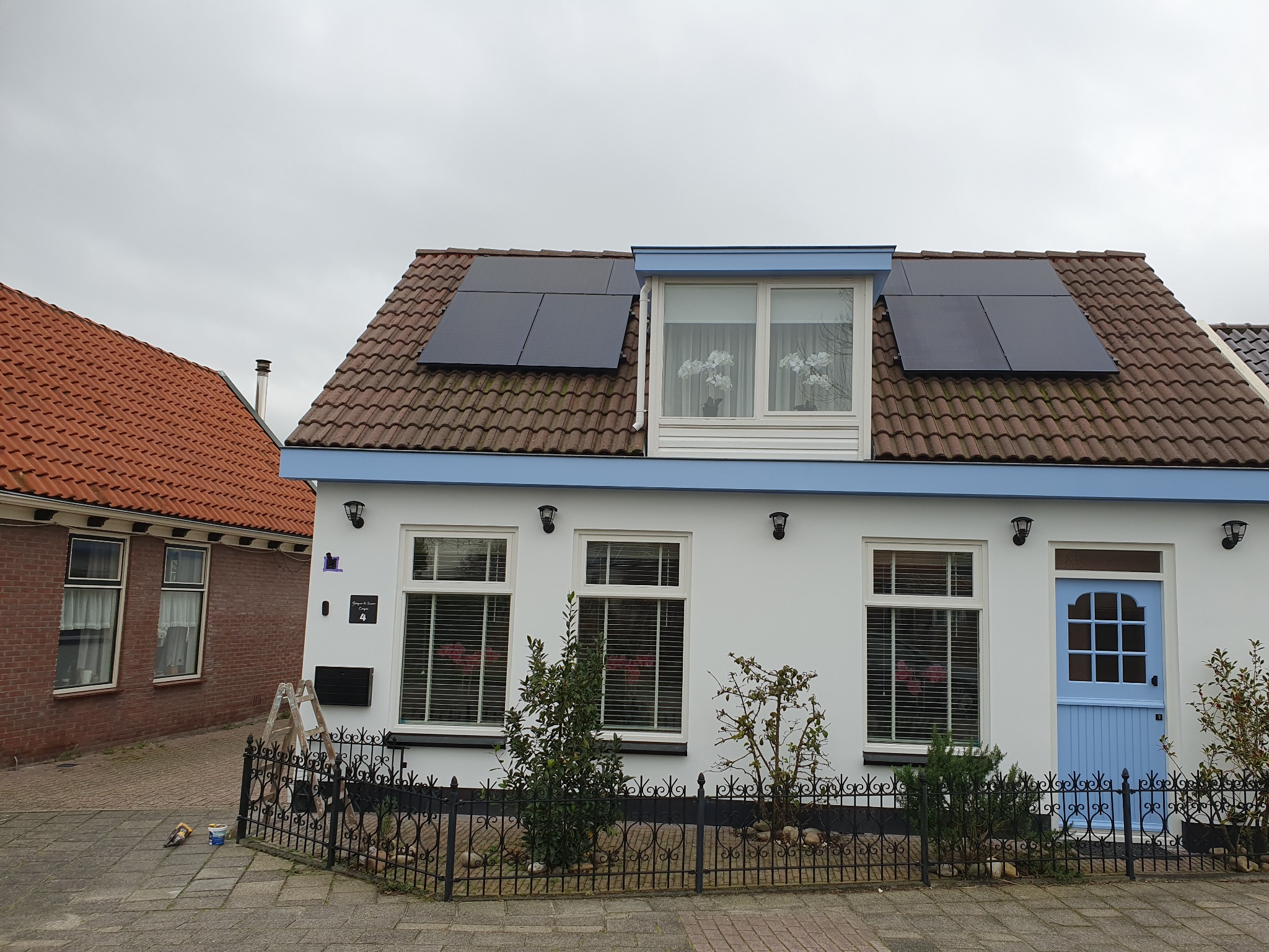Biały dom z niebieskimi akcentami i panelami słonecznymi na brązowym dachu, widziany z poziomu ulicy. W tle widoczny sąsiedni dom z czerwonej cegły.