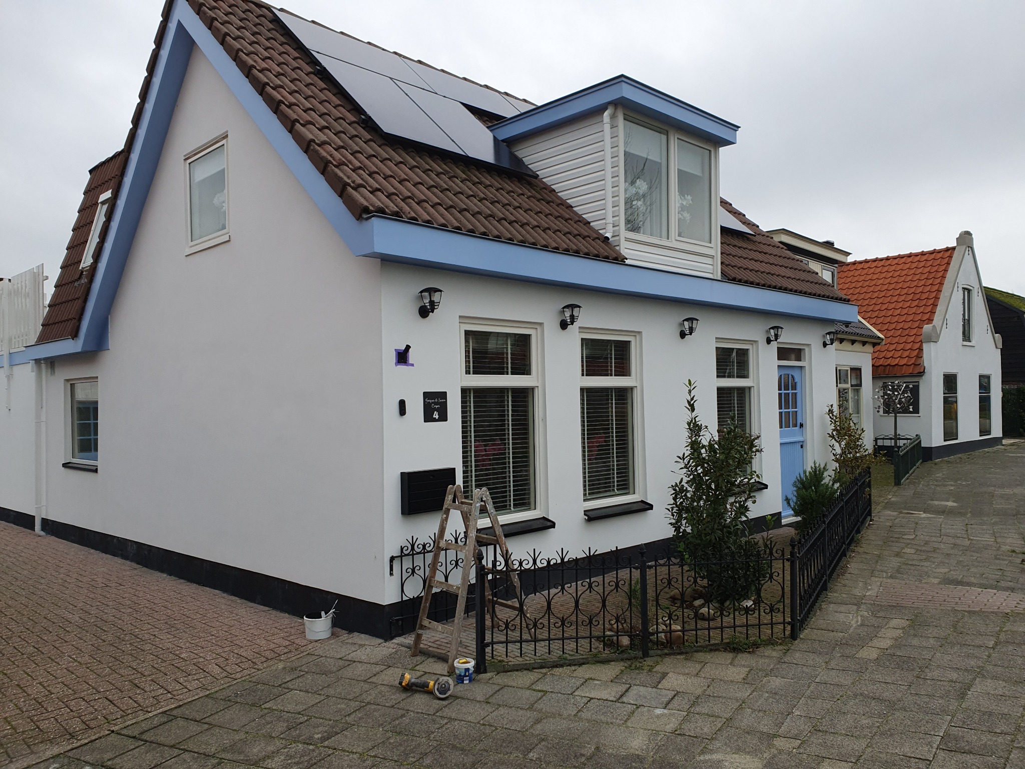 Biały dom z niebieskimi akcentami po renowacji elewacji w Brzegu. Widoczne panele słoneczne na dachu, drabina i akcesoria malarskie przy ścianie.