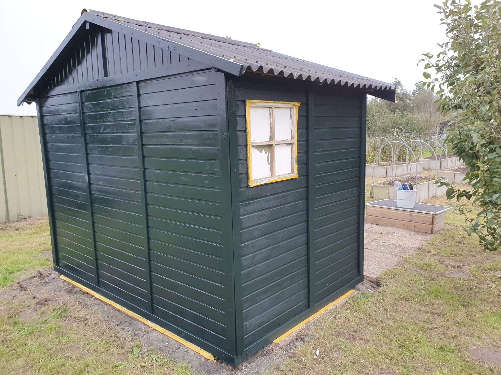 Pomazany zielenią domek narzędziowy z oknem, zabezpieczony taśmą malarską. Widoczne ślady świeżego malowania i elementy ogrodu w tle.