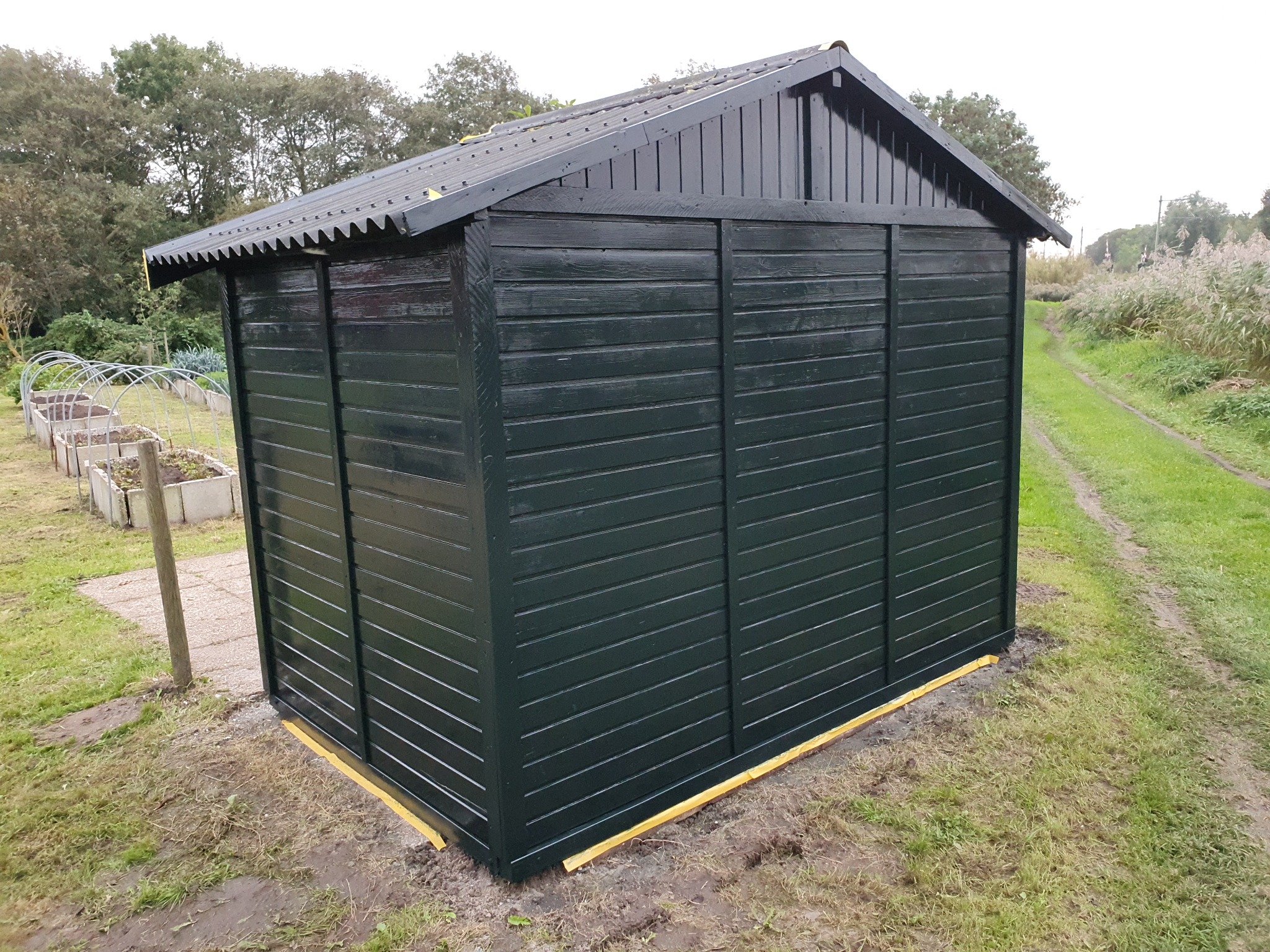 Ciemnozielona, świeżo pomalowana drewniana szopa ogrodowa z czarnym dachem, otoczona trawą. Żółta taśma malarska zabezpiecza dolną krawędź. Widoczne grządki warzywne w tle.