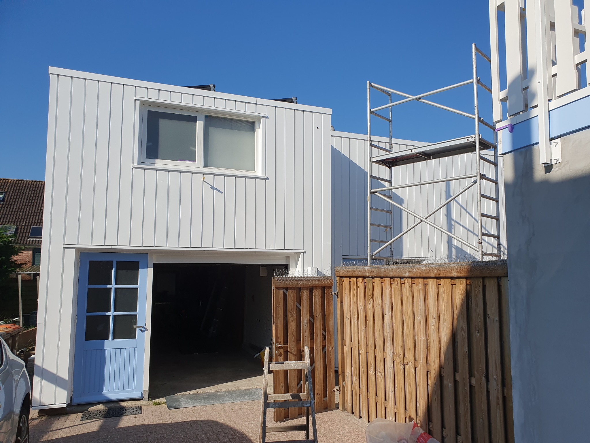 Biały budynek z błękitnymi drzwiami, widoczne rusztowanie i drewniane ogrodzenie. Całość w jasnym świetle słonecznym.