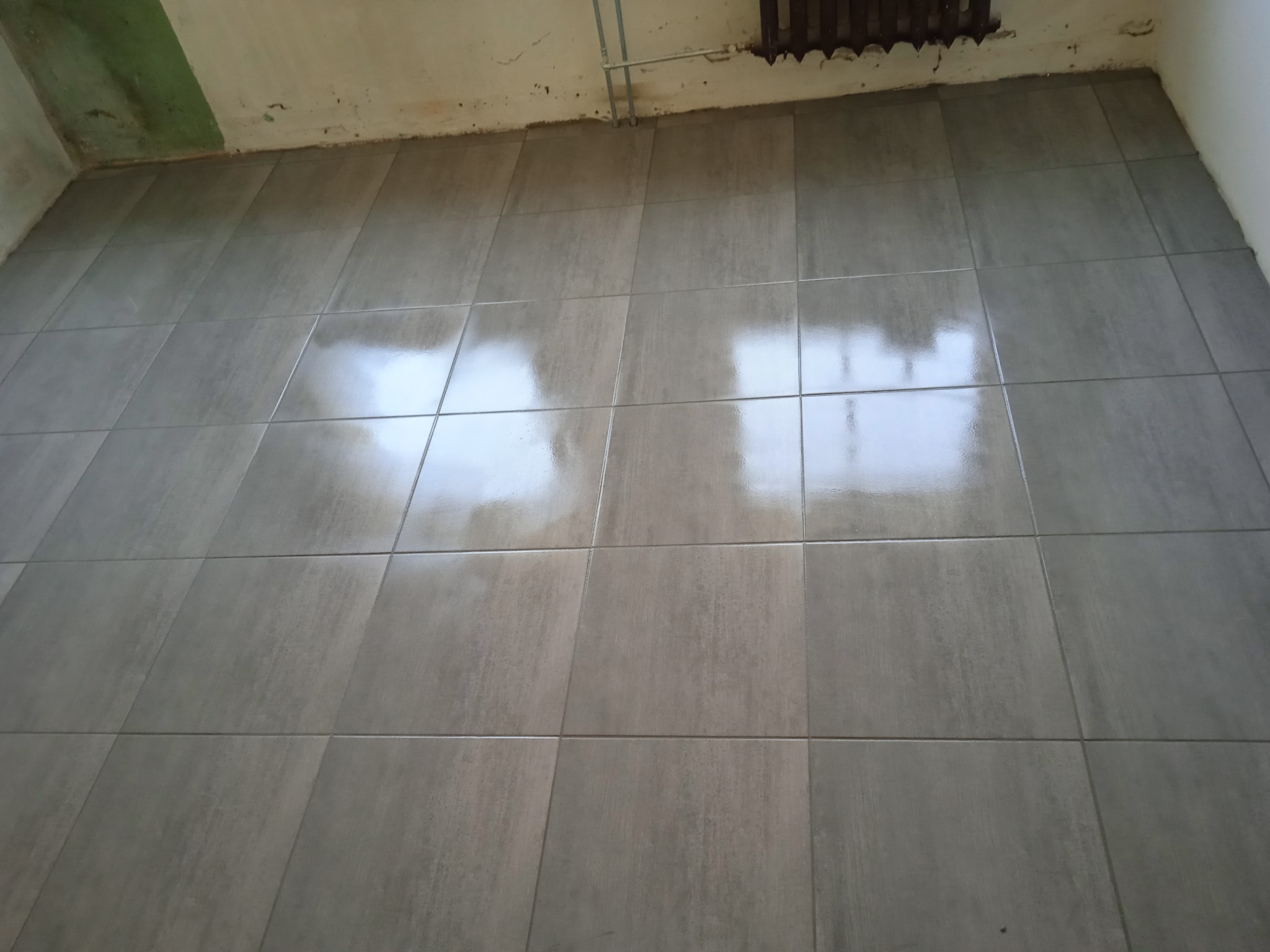 Ułożone, szare płytki podłogowe z widocznymi refleksami światła, nierówności ścian i rurka pod sufitem. Układ kwadratowy, fuga w kolorze płytek, surowy stan wykończenia.