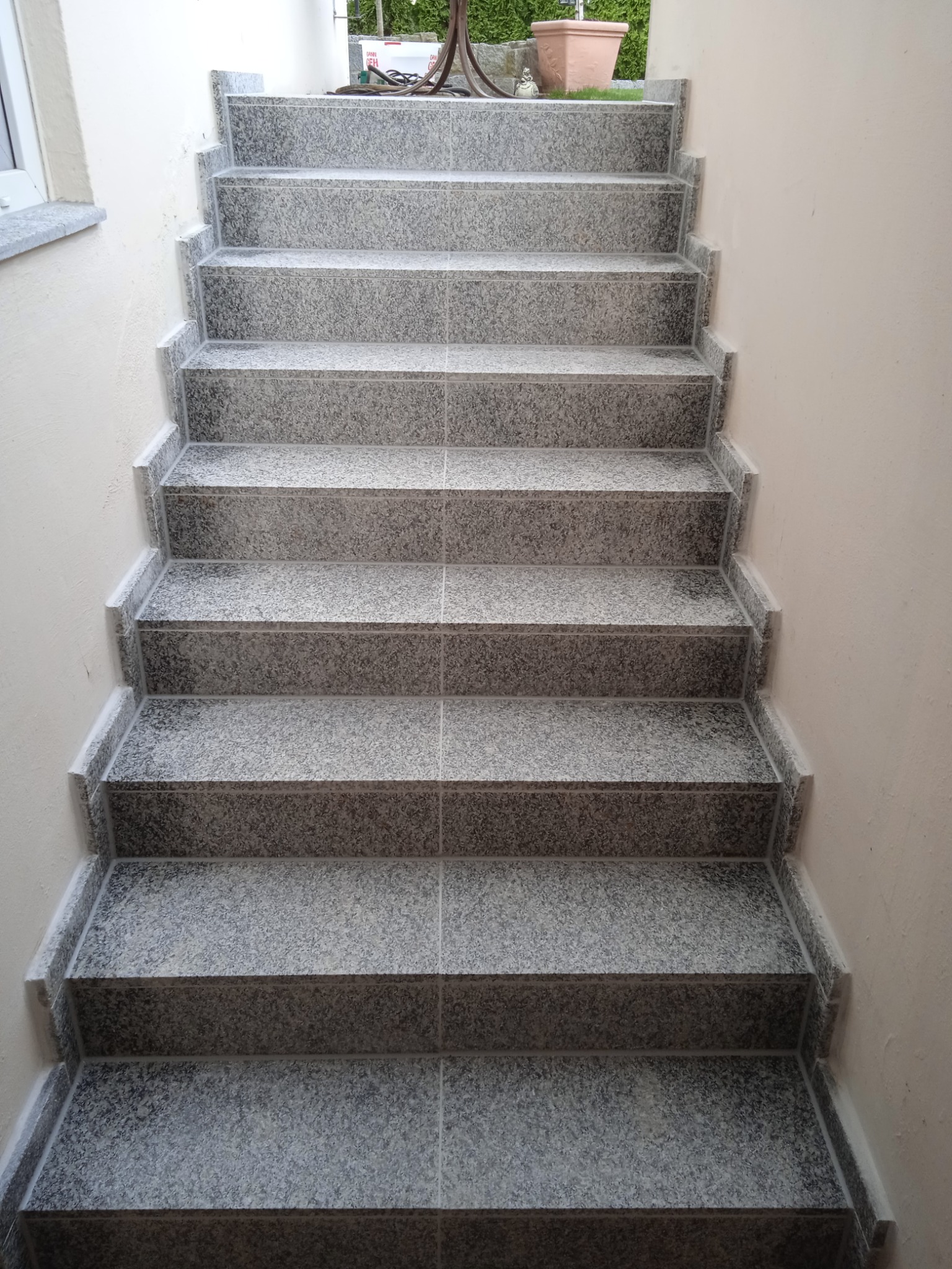 Wykończone schody zewnętrzne z szarego granitu, widok z dołu. Estetyczne połączenia płytek, jasne ściany, delikatne oświetlenie. Precyzyjne wykonanie.