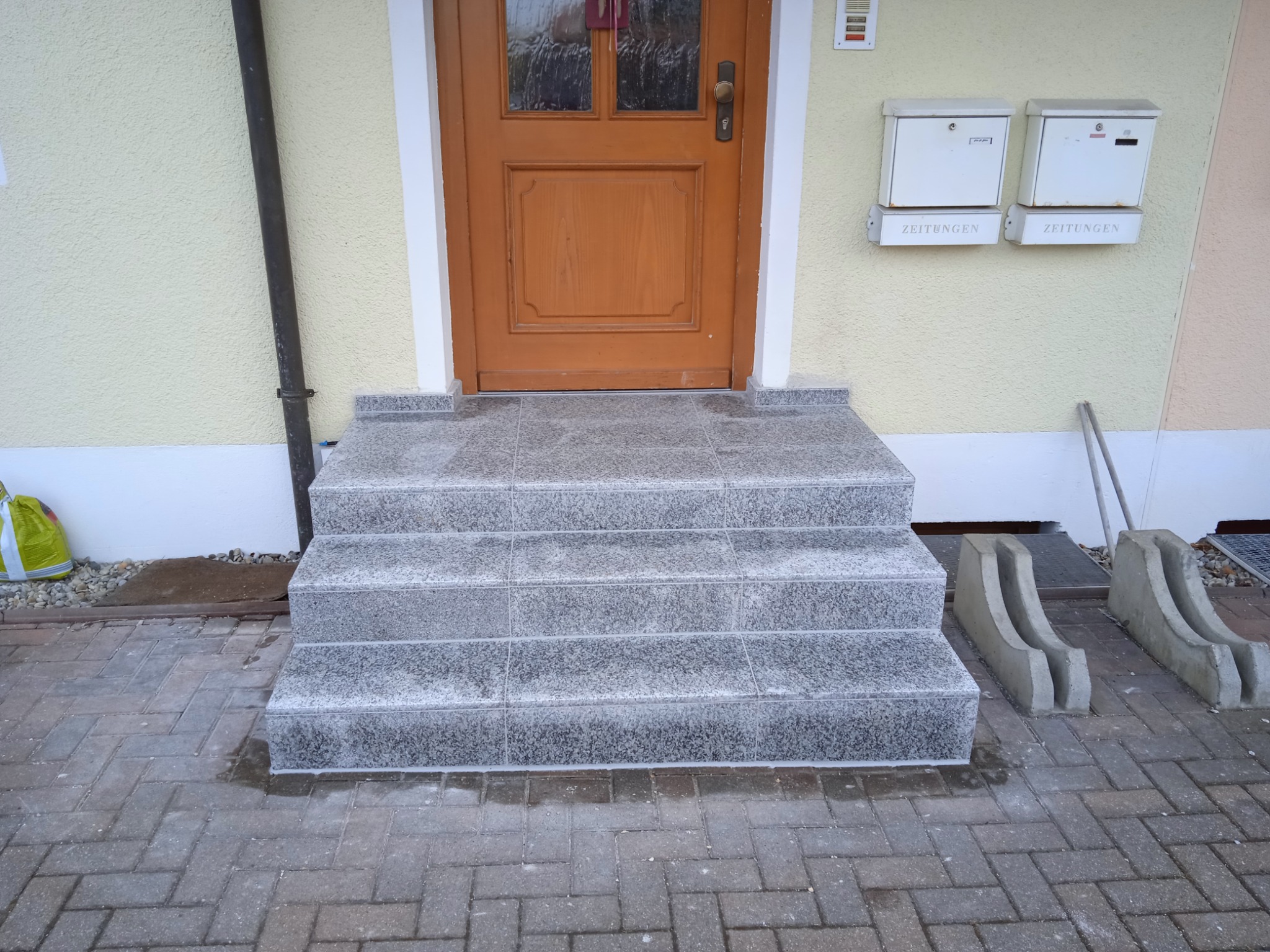 Granitowe schody wejściowe z szarą kostką brukową, prowadzące do drewnianych drzwi z szybami. Dwa białe pojemniki na gazety po prawej stronie. Ujęcie z dołu.