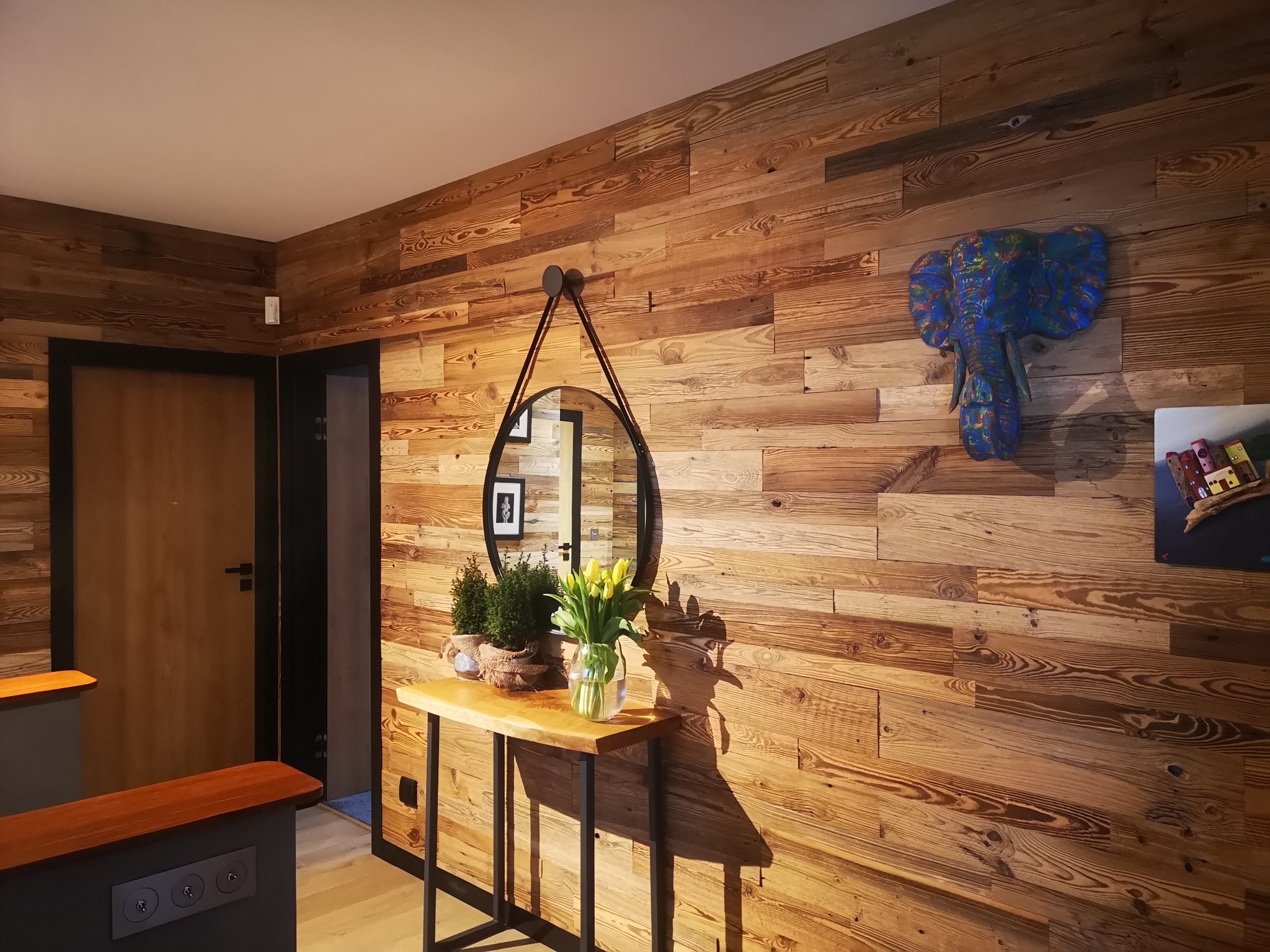 Drewniana ściana z poziomych paneli o wyraźnym usłojeniu, okrągłe lustro na skórzanym pasku, drewniany stolik z żółtymi tulipanami w wazonie i rzeźba głowy słonia w niebieskich barwach.