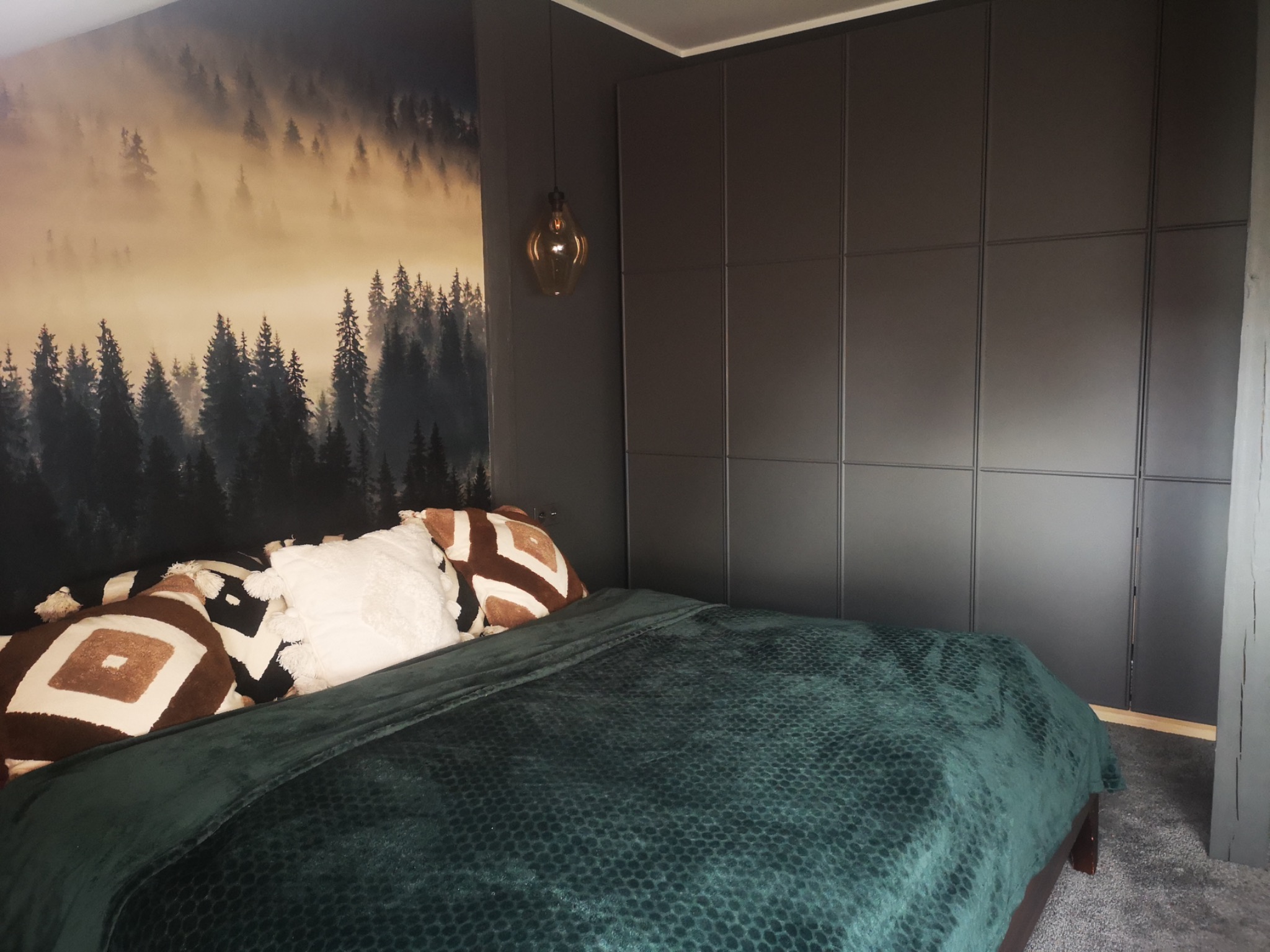 Nowoczesna sypialnia w ciemnych barwach z fototapetą przedstawiającą mglisty las, ciemnoszara zabudowa szafy na całą ścianę, łóżko z zieloną narzutą i dekoracyjnymi poduszkami w stylu boho, wisząca...