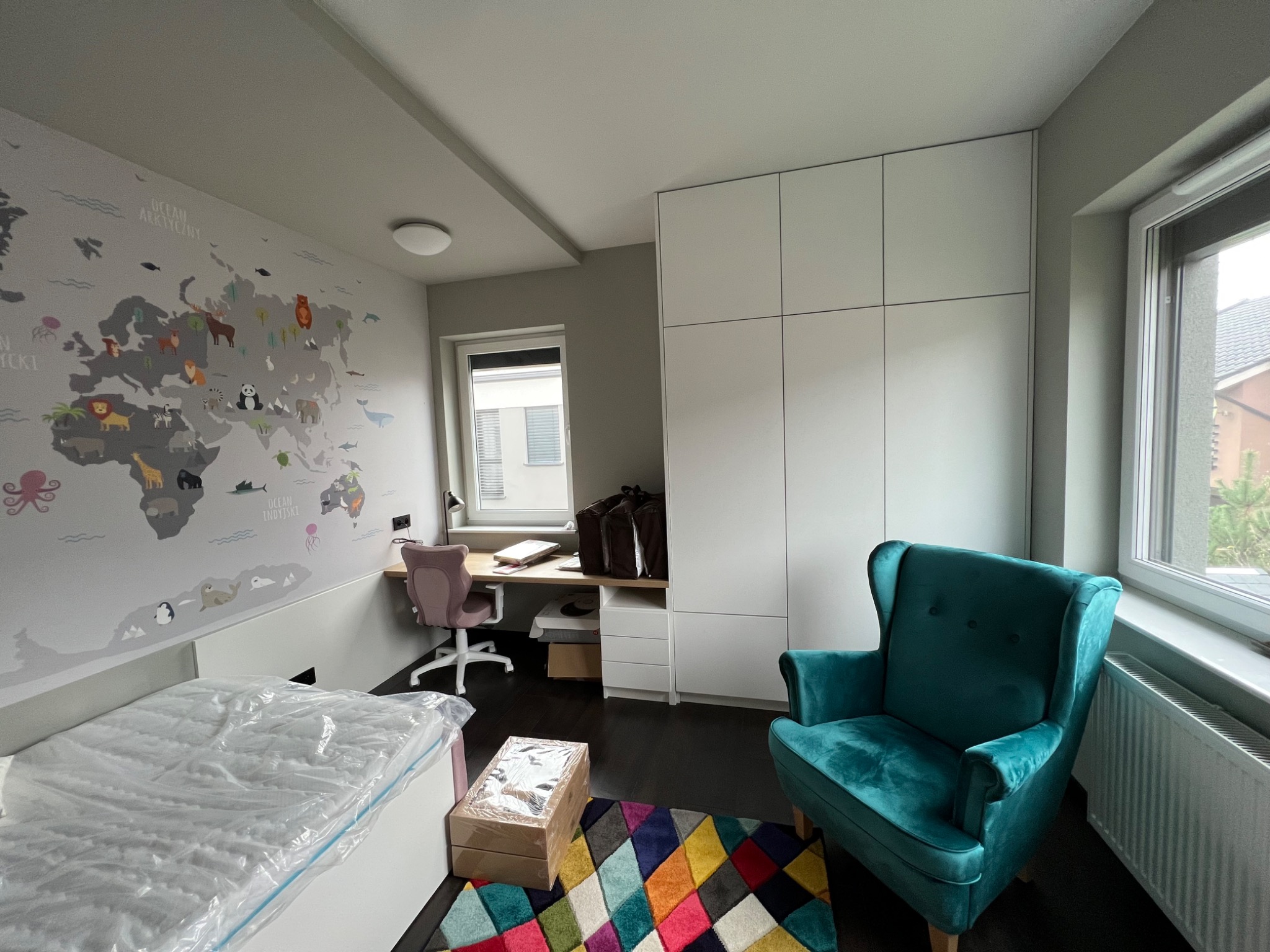 Pokój dziecięcy z mapą świata na ścianie, biurkiem, krzesłem, turkusowym fotelem i szafą w zabudowie, z nowym dywanem w geometryczne wzory i zapakowanym materacem na łóżku.