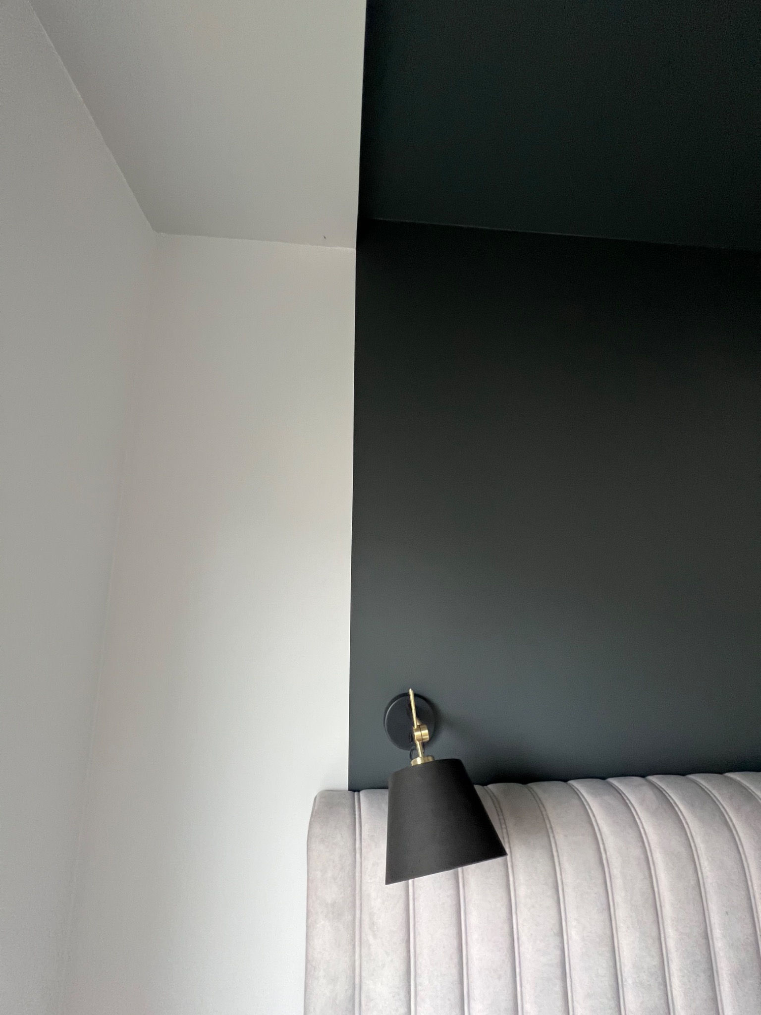 Ściana pomalowana dwukolorowo - biały i grafitowy, z czarną lampką nocną z elementami w kolorze złota nad wezgłowiem łóżka w odcieniach szarości z pionowymi przeszyciami.