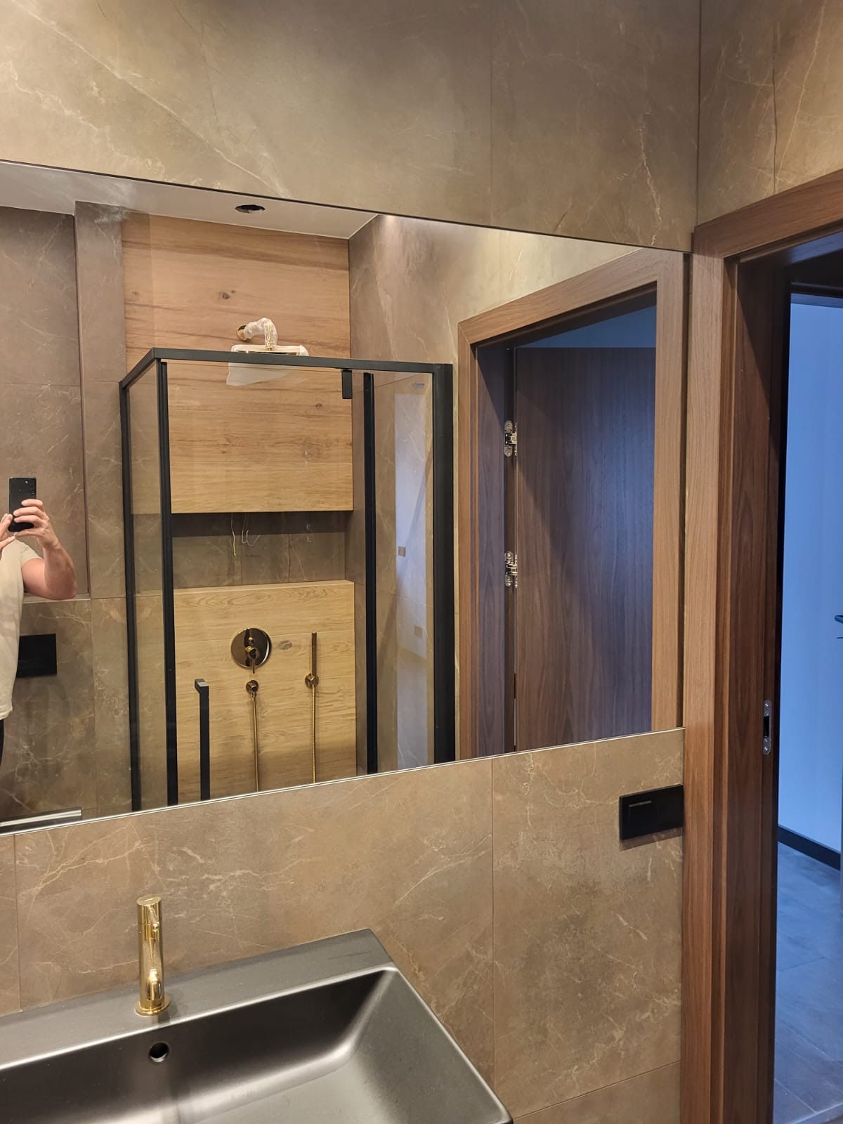 Nowoczesna łazienka w odcieniach beżu i brązu, z kabiną prysznicową z elementami drewnopodobnymi i czarnymi ramami, widoczna w lustrze nad umywalką z złotą baterią.