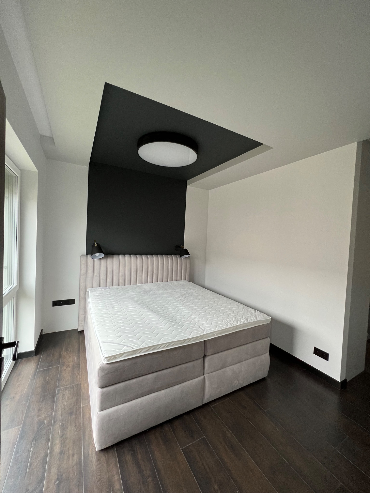 Sypialnia z łóżkiem kontynentalnym, ciemną wnęką nad wezgłowiem i okrągłą lampą sufitową, parkiet w odcieniach brązu.