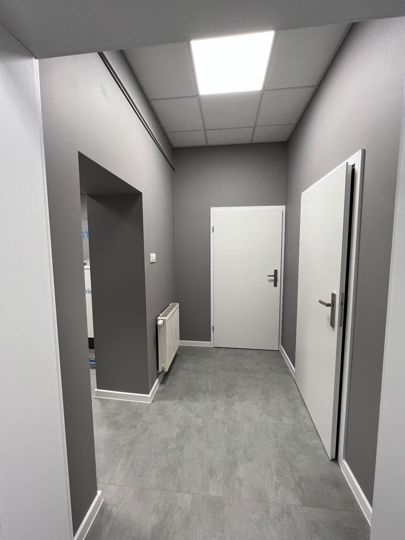 Wnętrze korytarza z szarą ścianą, białymi drzwiami i podłogą z szarych płytek, widoczny biały grzejnik i sufit kasetonowy z oświetleniem LED.