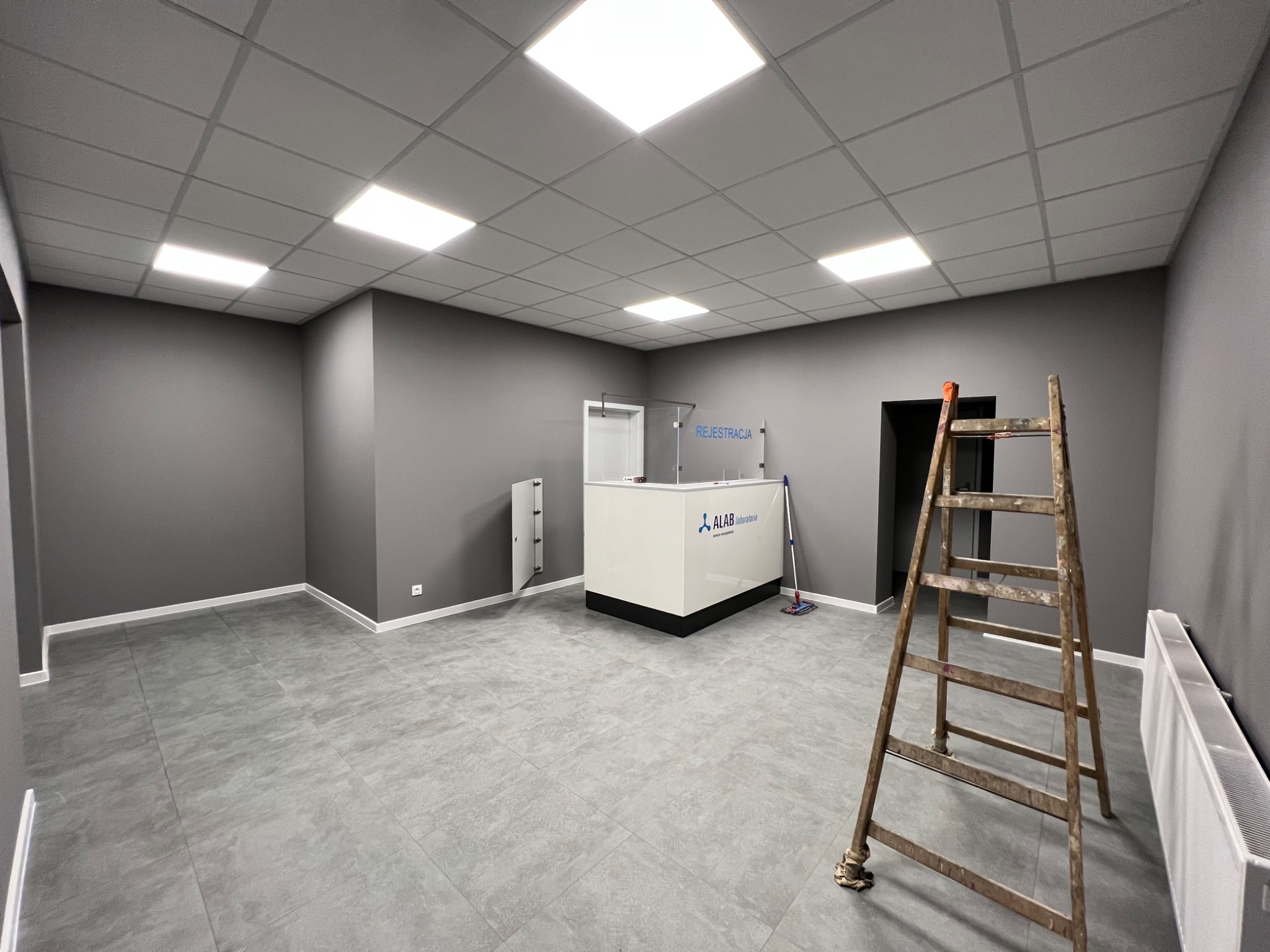 Szare wnętrze biurowe z recepcją ALAB Laboratoria, widoczna drewniana drabina, płytki podłogowe imitujące kamień, sufit podwieszany z kwadratowymi lampami.