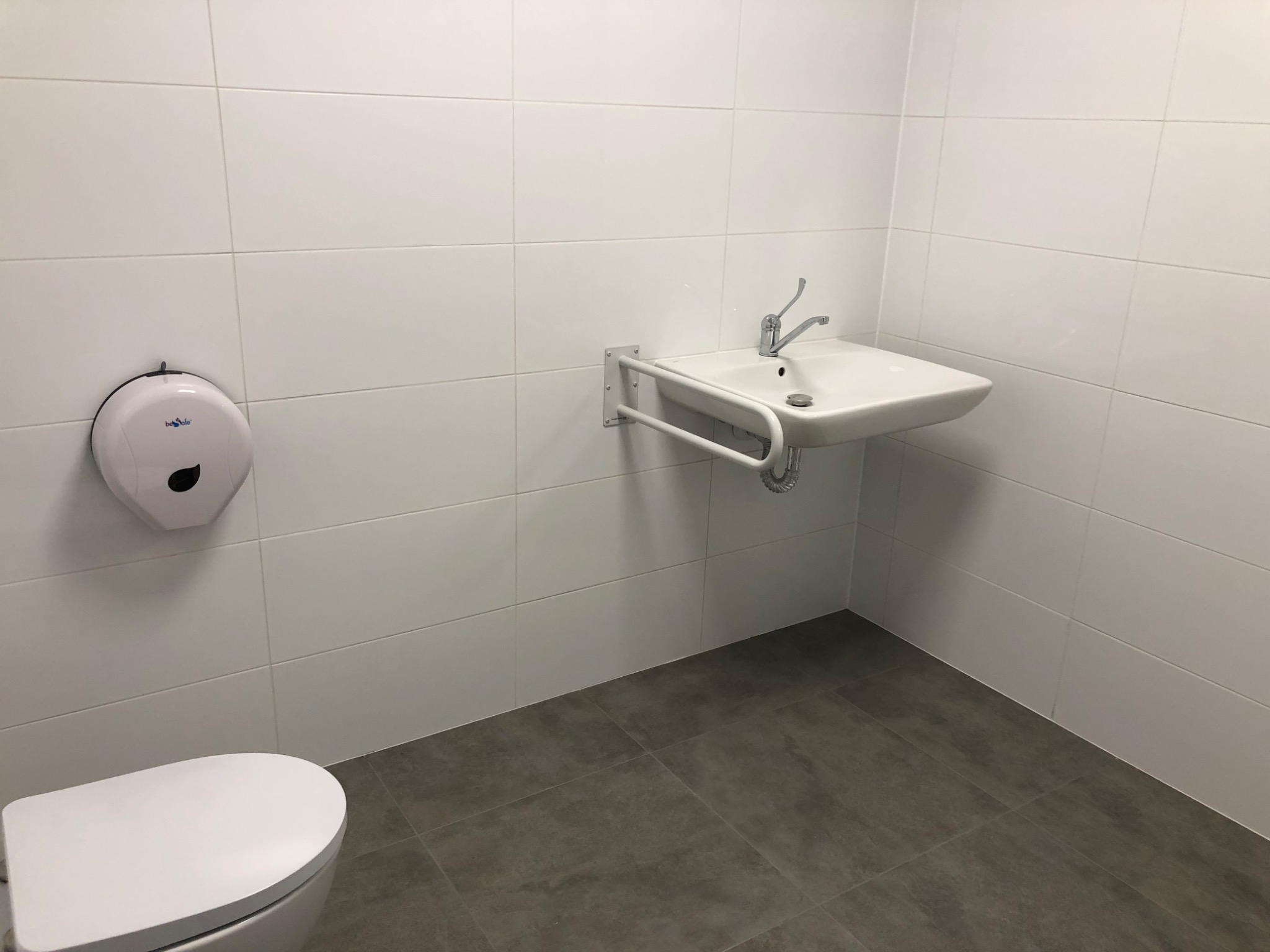 Łazienka dla niepełnosprawnych z białą umywalką z uchwytem, białą toaletą i dozownikiem ręczników na ścianie z białych płytek, szara podłoga z płytek.