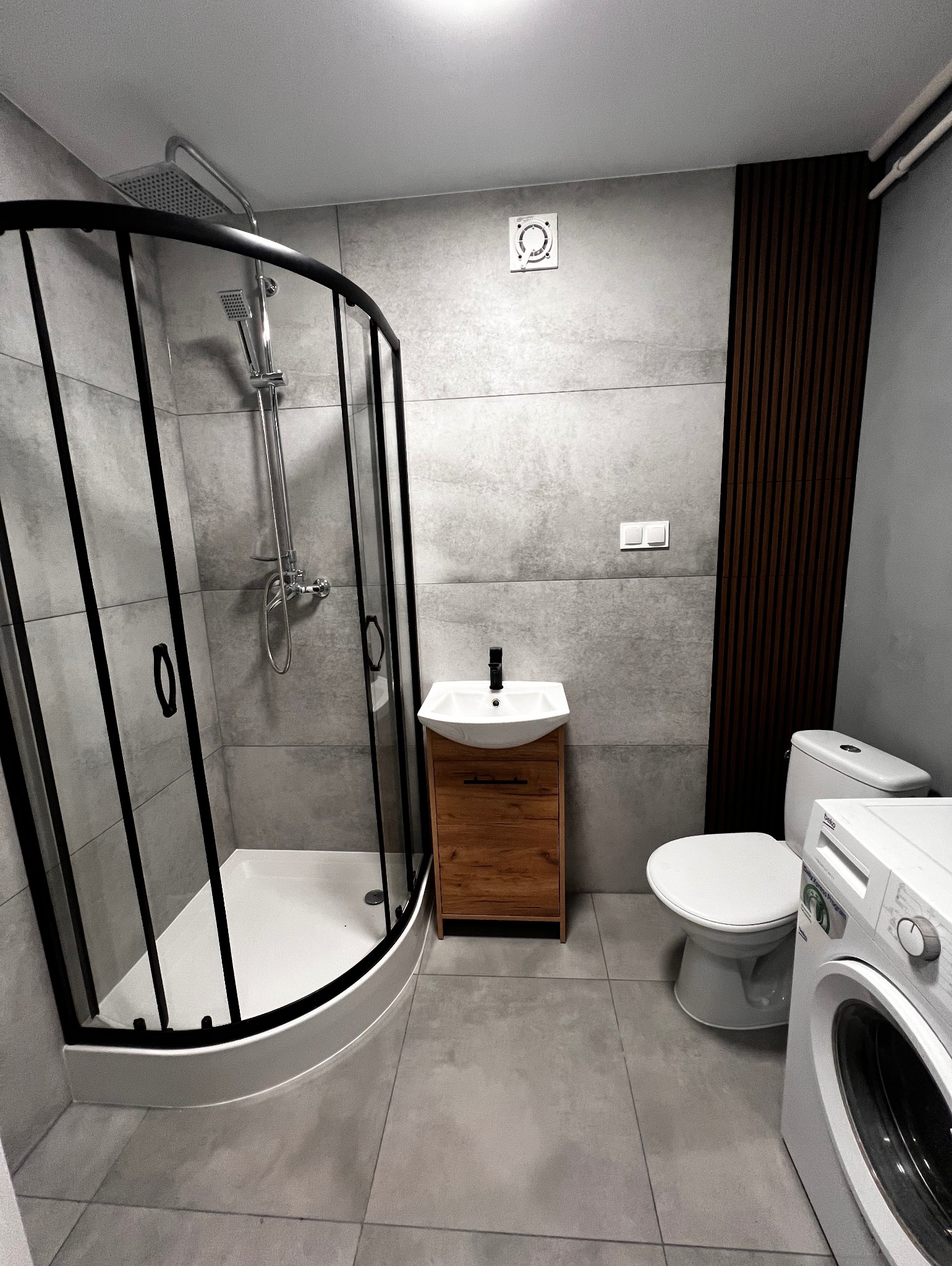 Nowoczesna łazienka w szarościach z kabiną prysznicową z czarnymi profilami, umywalką z drewnianą szafką, toaletą i pralką.