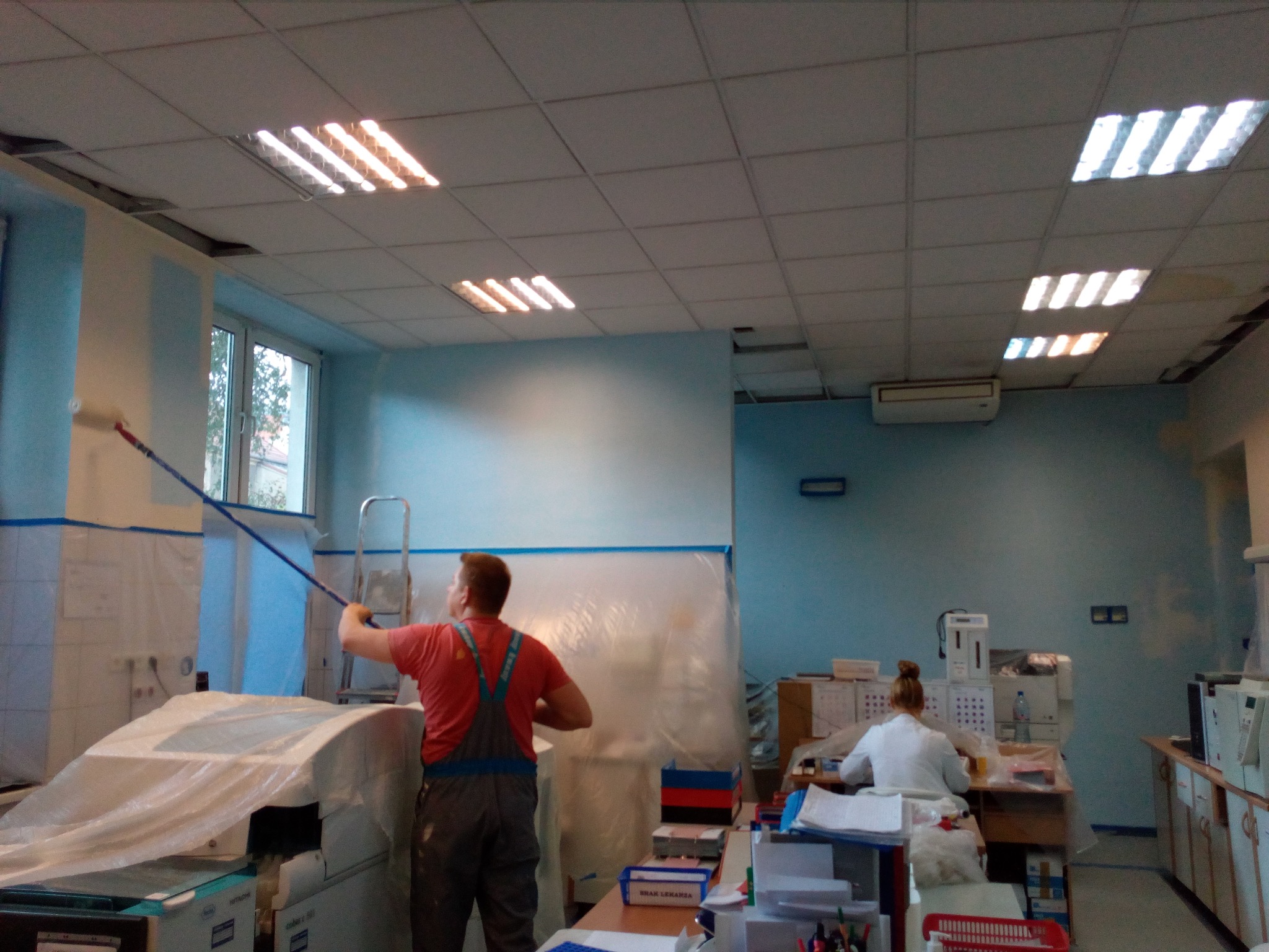 Mężczyzna w kombinezonie maluje ścianę w pomieszczeniu biurowym na jasny niebieski kolor za pomocą wałka na długim kiju, w tle biurko z pracownikiem i sprzętem biurowym, podłoga zabezpieczona folią.