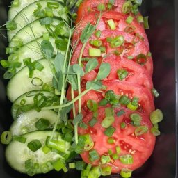 Catering do pełna - Kompozycja z plastrów ogórka i pomidora, posypana szczypiorkiem i udekorowana pędami groszku, ułożona w czarnym naczyniu.