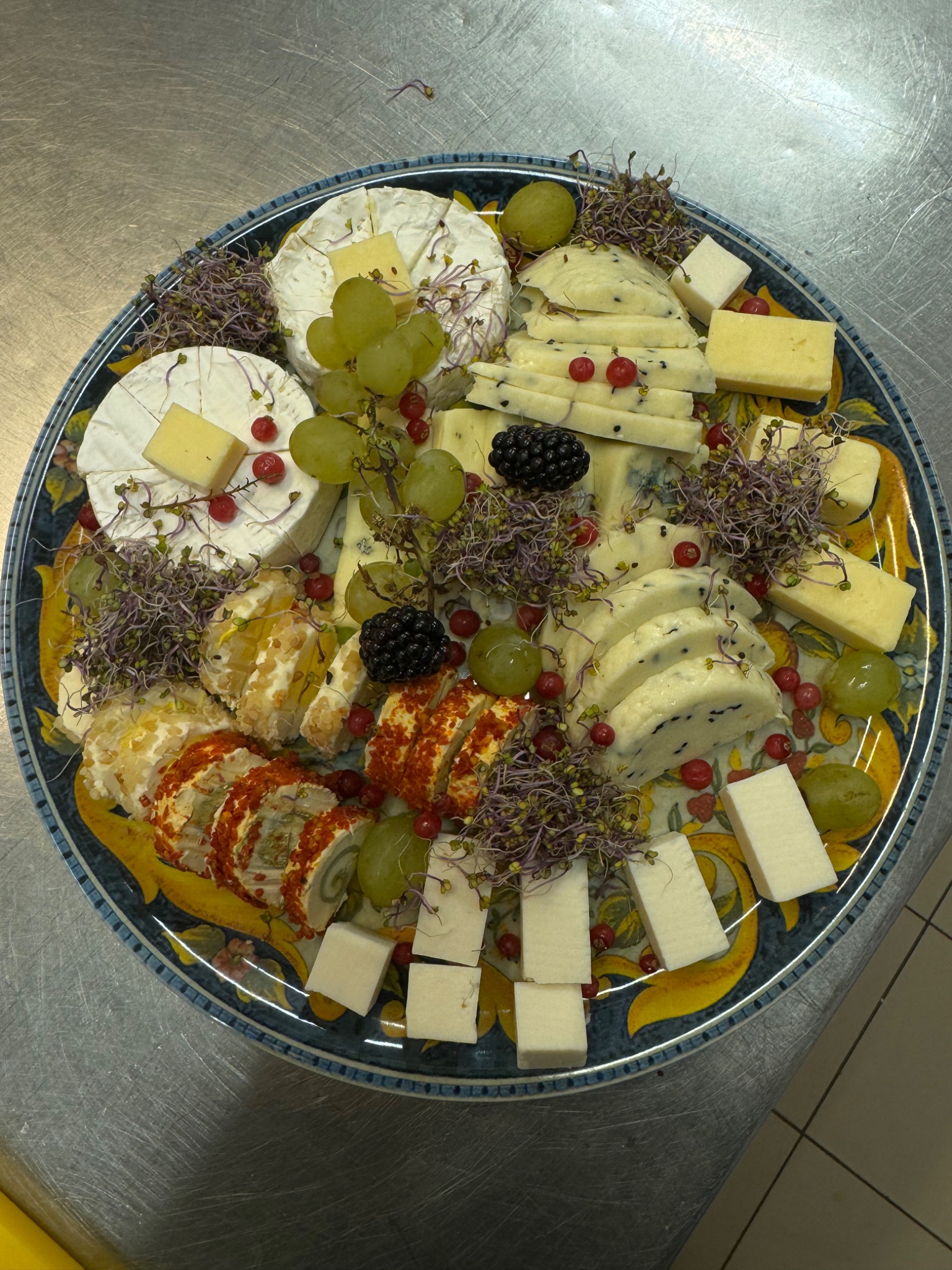 Dekoracyjna taca z różnorodnym wyborem serów, winogron, jeżyn i czerwonych porzeczek, ozdobiona mikrolistkami, prezentowana na metalowym blacie.