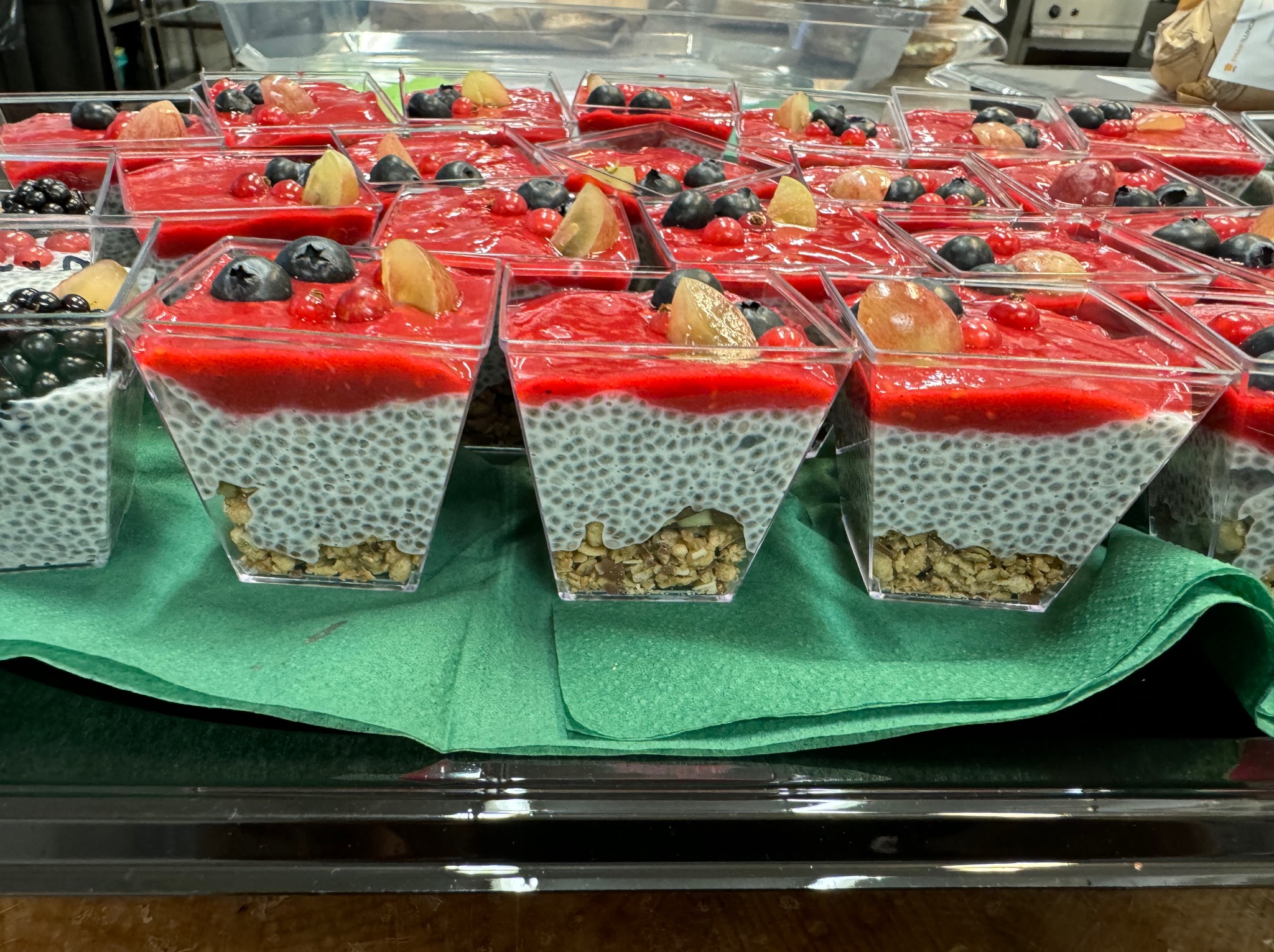 Deser chia z musem truskawkowym, borówkami, czerwoną porzeczką i physalis w przezroczystych, kwadratowych naczyniach, ułożony na zielonej serwetce.