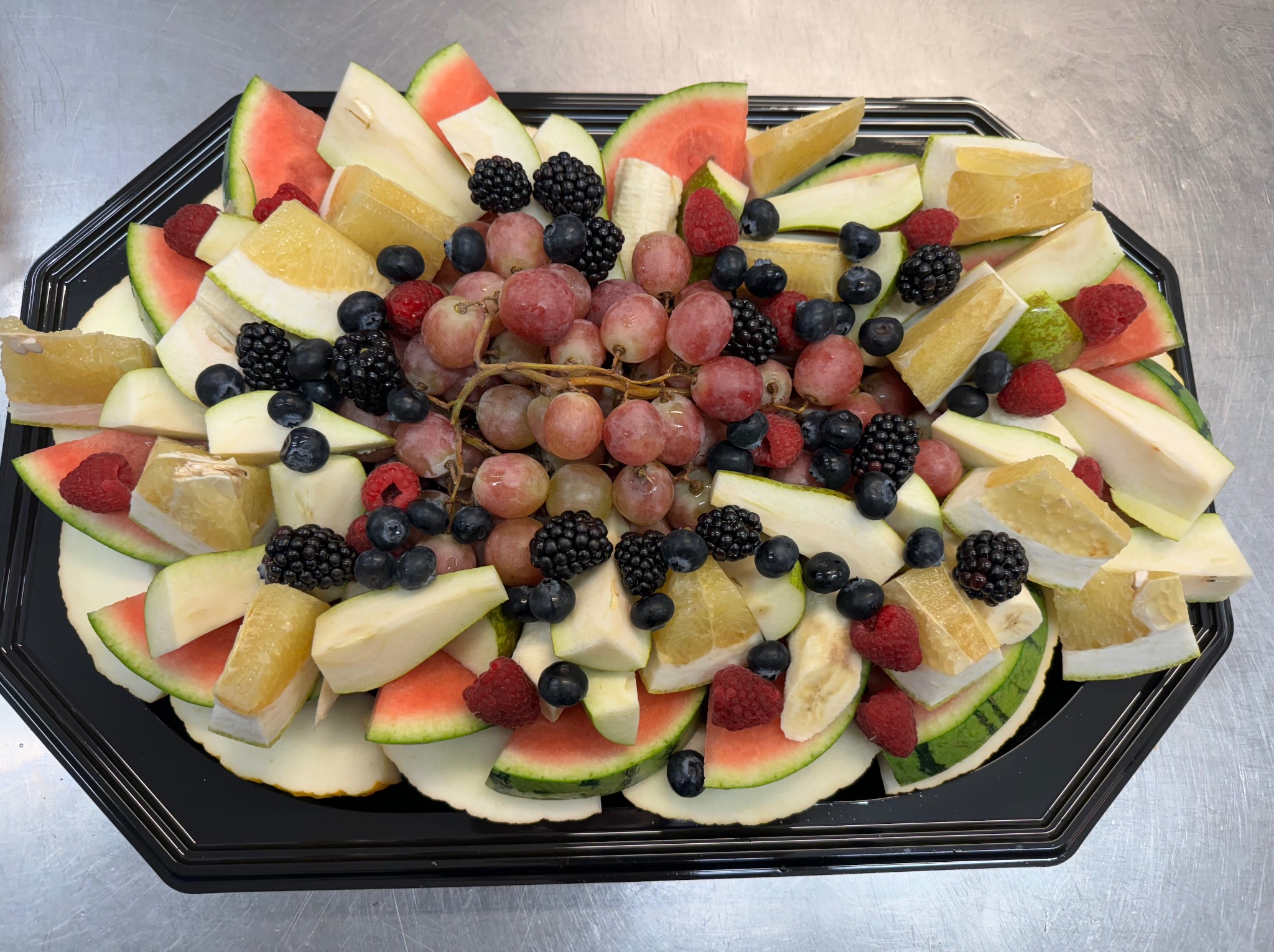 Dekoracyjna czarna taca z różnorodnymi pokrojonymi owocami: arbuz, melon, grejpfrut, gruszka, maliny, jeżyny, borówki i winogrona.