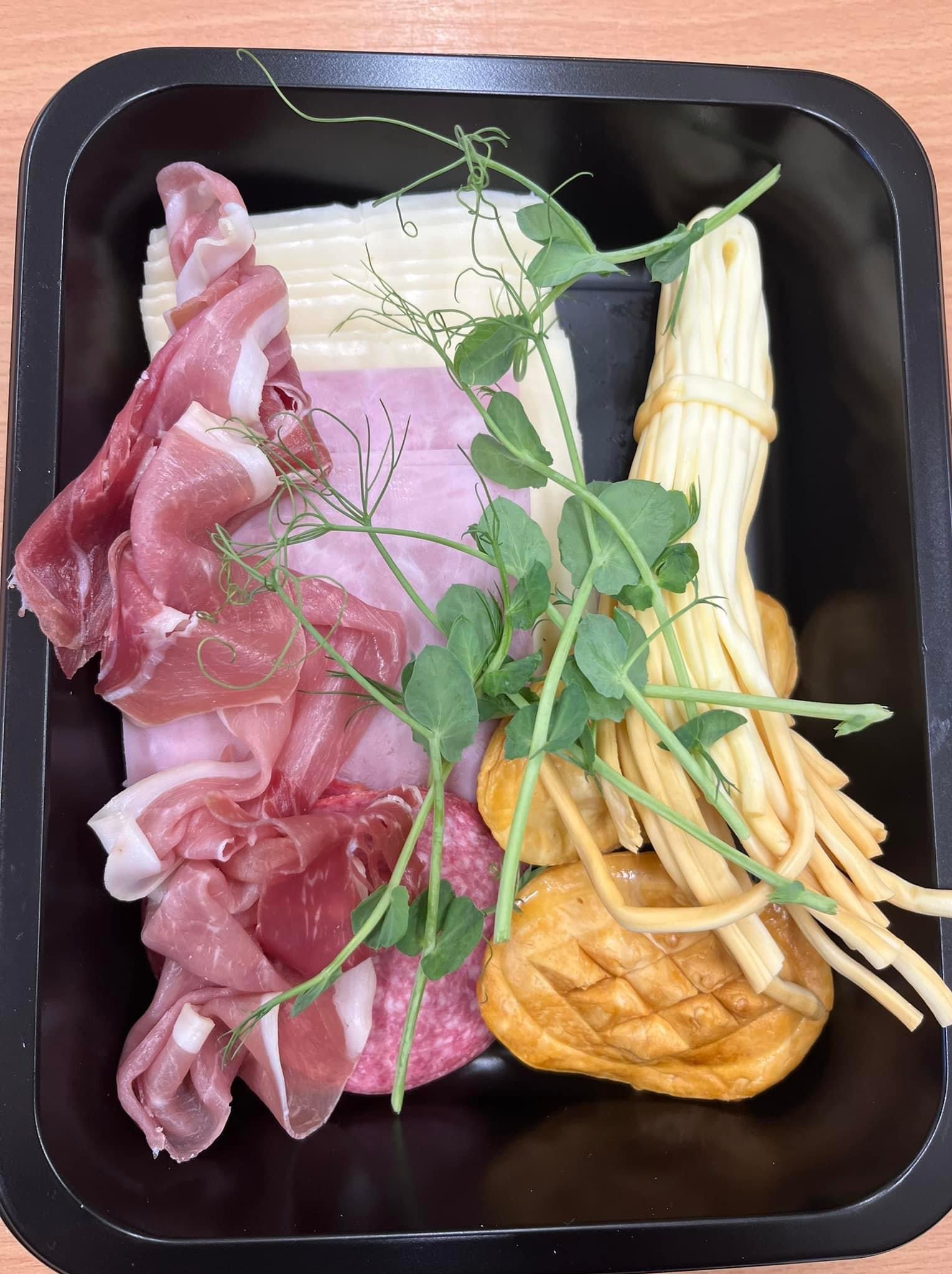 Czarna tacka z przekąskami: plasterki szynki parmeńskiej, salami, ser żółty, ser wędzony w warkoczach oraz dekoracja z pędów groszku.