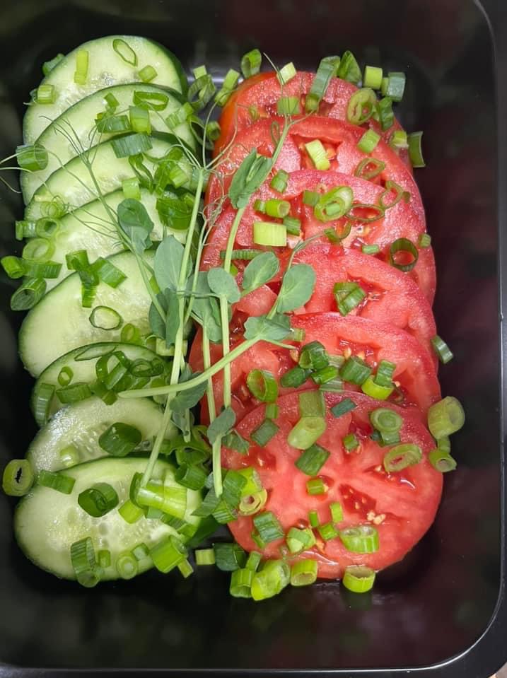 Kompozycja z plastrów ogórka i pomidora, posypana szczypiorkiem i udekorowana pędami groszku, ułożona w czarnym naczyniu.