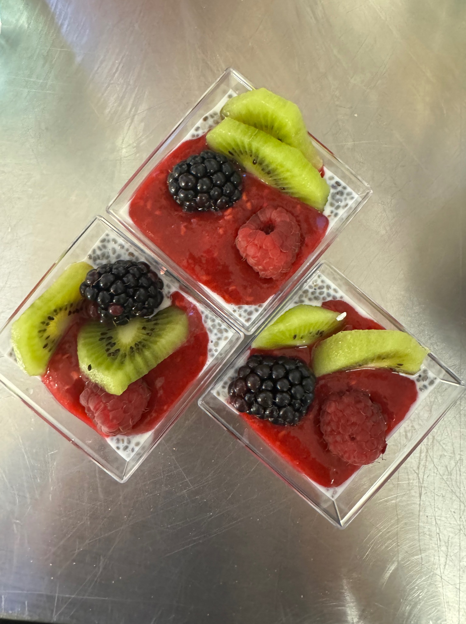 Trzy kwadratowe, przezroczyste miseczki z deserem chia z musem malinowym, jeżynami i kiwi na metalowym stole. Deser udekorowany pojedynczą maliną. Widoczne białe nasiona chia.