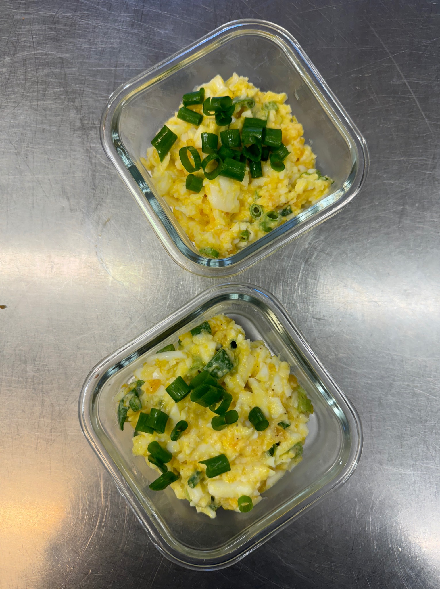 Dwie szklane miseczki z sałatką jajeczną, posypaną szczypiorkiem, ustawione na metalowej powierzchni.