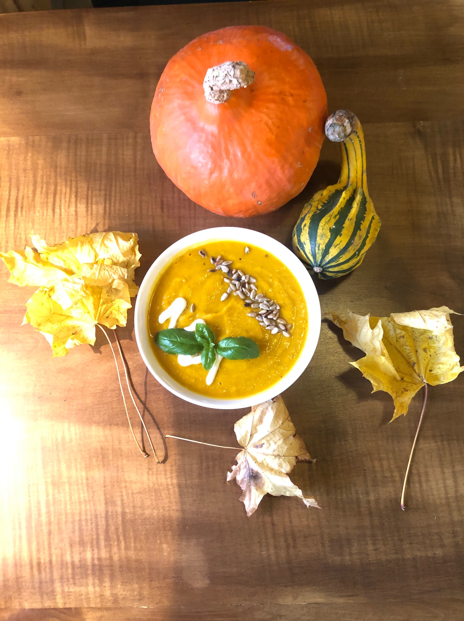 Krem z dyni w białej miseczce, udekorowany śmietaną, bazylią i prażonymi pestkami słonecznika, otoczony dynią, ozdobną tykwą i opadłymi liśćmi klonu na drewnianym stole.