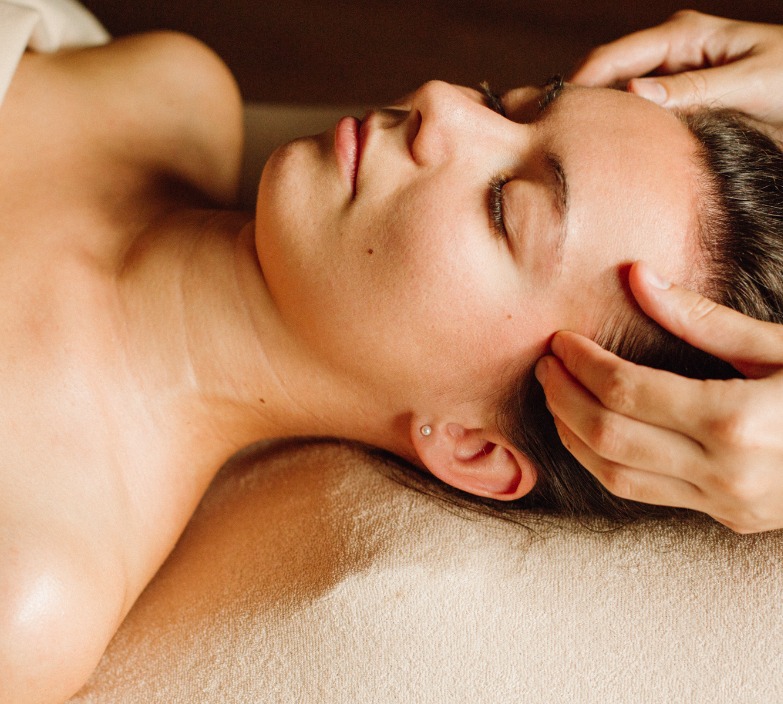 Kobieta z zamkniętymi oczami podczas masażu głowy, widoczny kolczyk w uchu i delikatne oświetlenie.