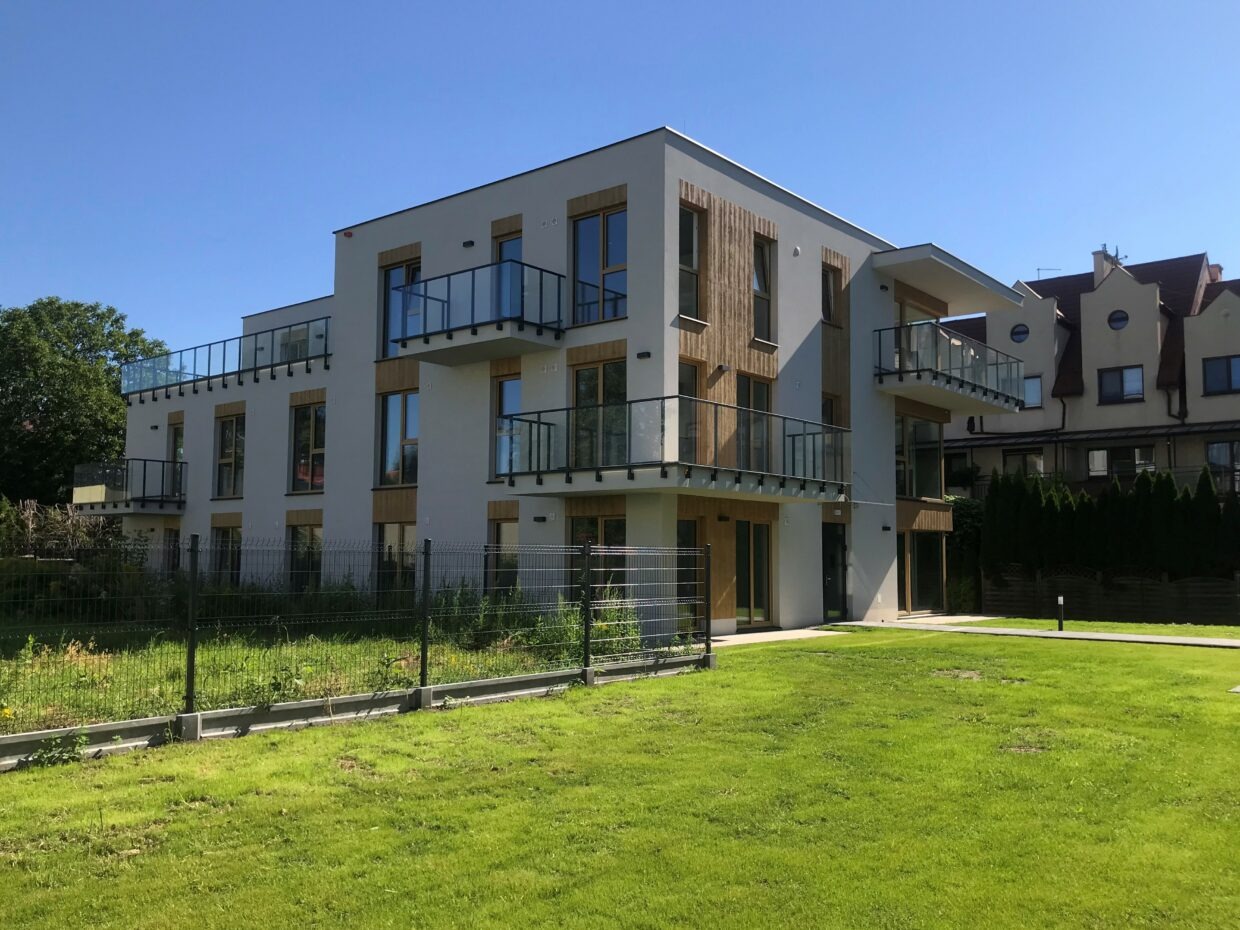 Nowoczesny budynek mieszkalny z balkonami i elementami drewnianymi, otoczony trawnikiem i ogrodzeniem z siatki.